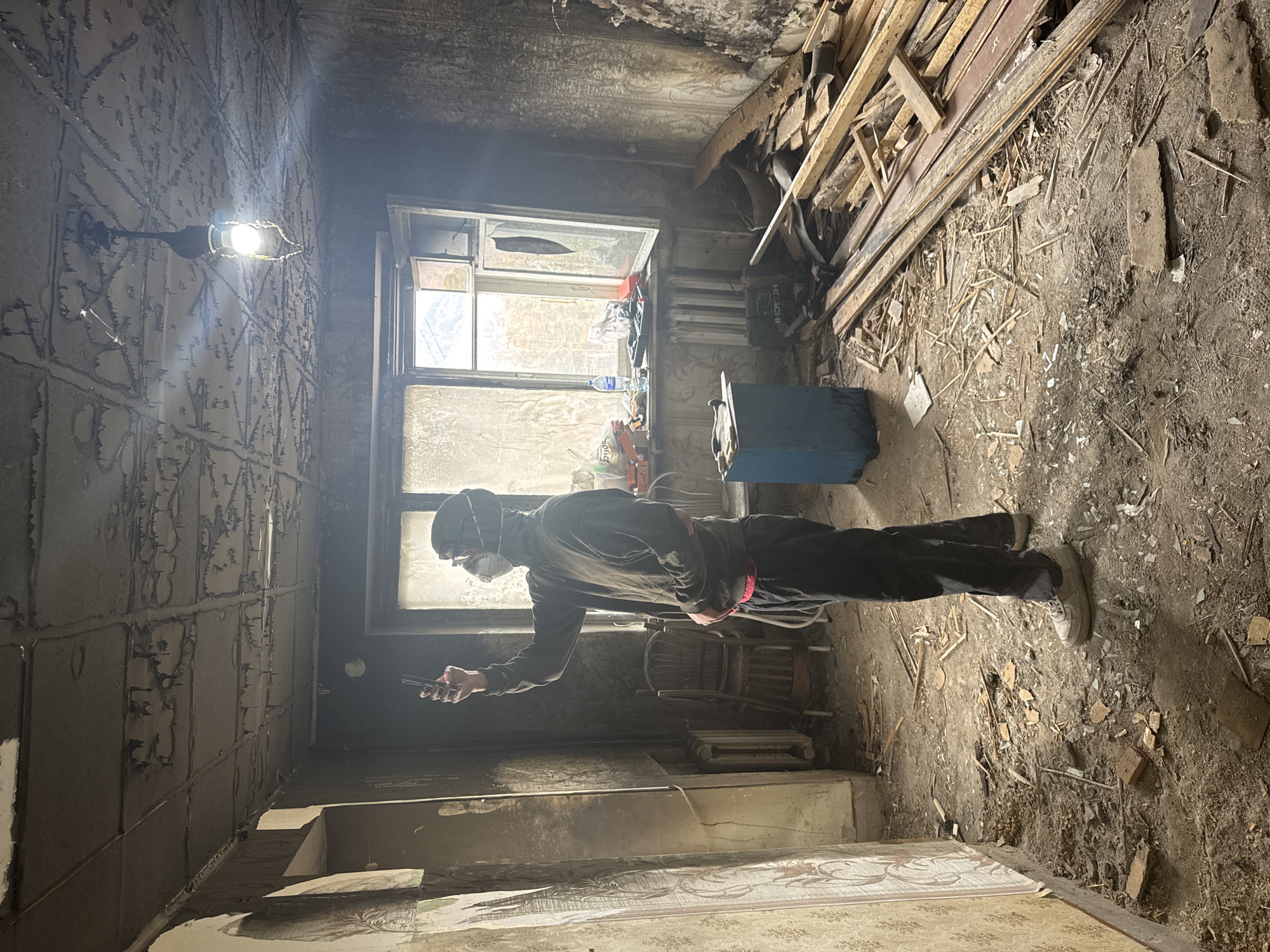 A man wearing a face mask and dark clothing stands in a severely damaged room, possibly a residential interior. He holds a cell phone in his hand, as if taking a picture or recording. The room shows signs of fire damage and neglect, with soot-covered walls, broken windows, and debris scattered on the floor. Piles of wooden planks and construction materials are stacked against one wall. A light bulb hangs bare from the ceiling, providing stark illumination to the scene. A partially visible chair and radiator stand near the window. The overall impression is one of dereliction and destruction. It is claimed this location is in Divnogorsk, Russia.