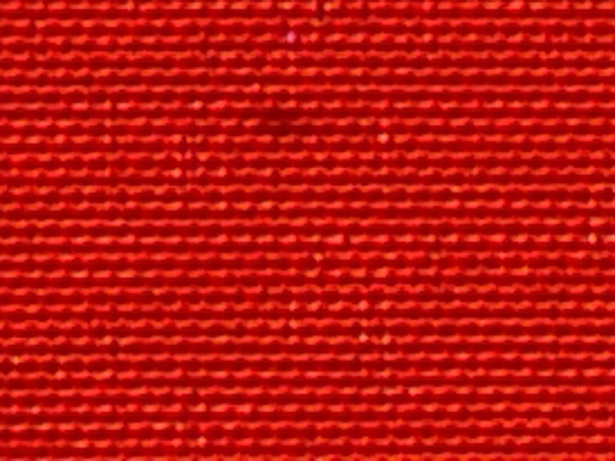 The image is a close-up, abstract shot of a bright red, woven material. The weave creates a textured pattern of vertical lines with subtle horizontal interruptions. The texture appears to be consistent throughout the frame, with no discernible objects, people, or environmental context visible. The lighting creates highlights and shadows that emphasize the texture of the fabric. There is no information in the image that could be used to determine a location in Orenburg, Russia.
