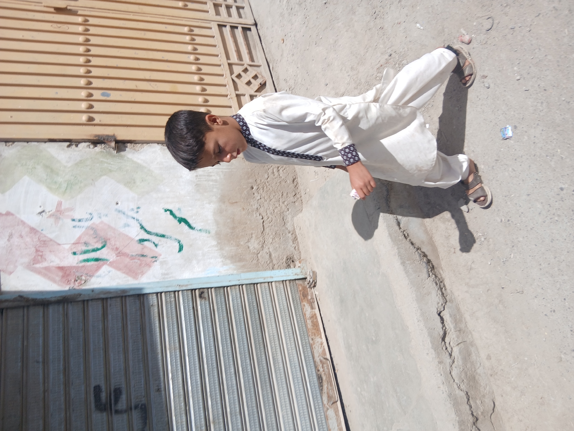 A young person in traditional Pakistani clothing walks on a sidewalk in Quetta City Tehsil, Pakistan. The person is wearing a white shalwar kameez with black patterned trim and brown sandals. The sidewalk is concrete and cracked. A metal roll-up door and a wall with graffiti are to the left. A corrugated metal door is visible above the wall.