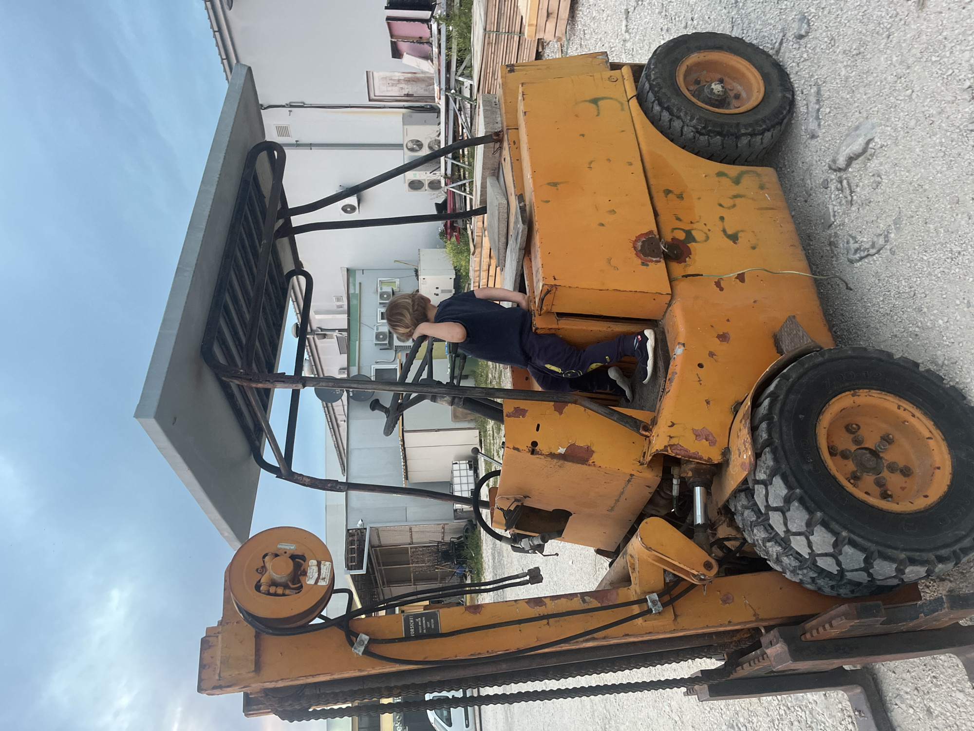 The image captures a young boy standing near an old, orange forklift in an outdoor environment. The location appears to be an industrial area in the city of Unknown, Unknown, with a building featuring multiple air conditioning units in the background. The forklift is stationary, with worn tires and visible signs of rust and age. The boy is wearing a dark shirt and pants and seems to be peering into the driver's area of the forklift. Additional details include lumber and other materials stacked near the building. The ground is covered with gravel.