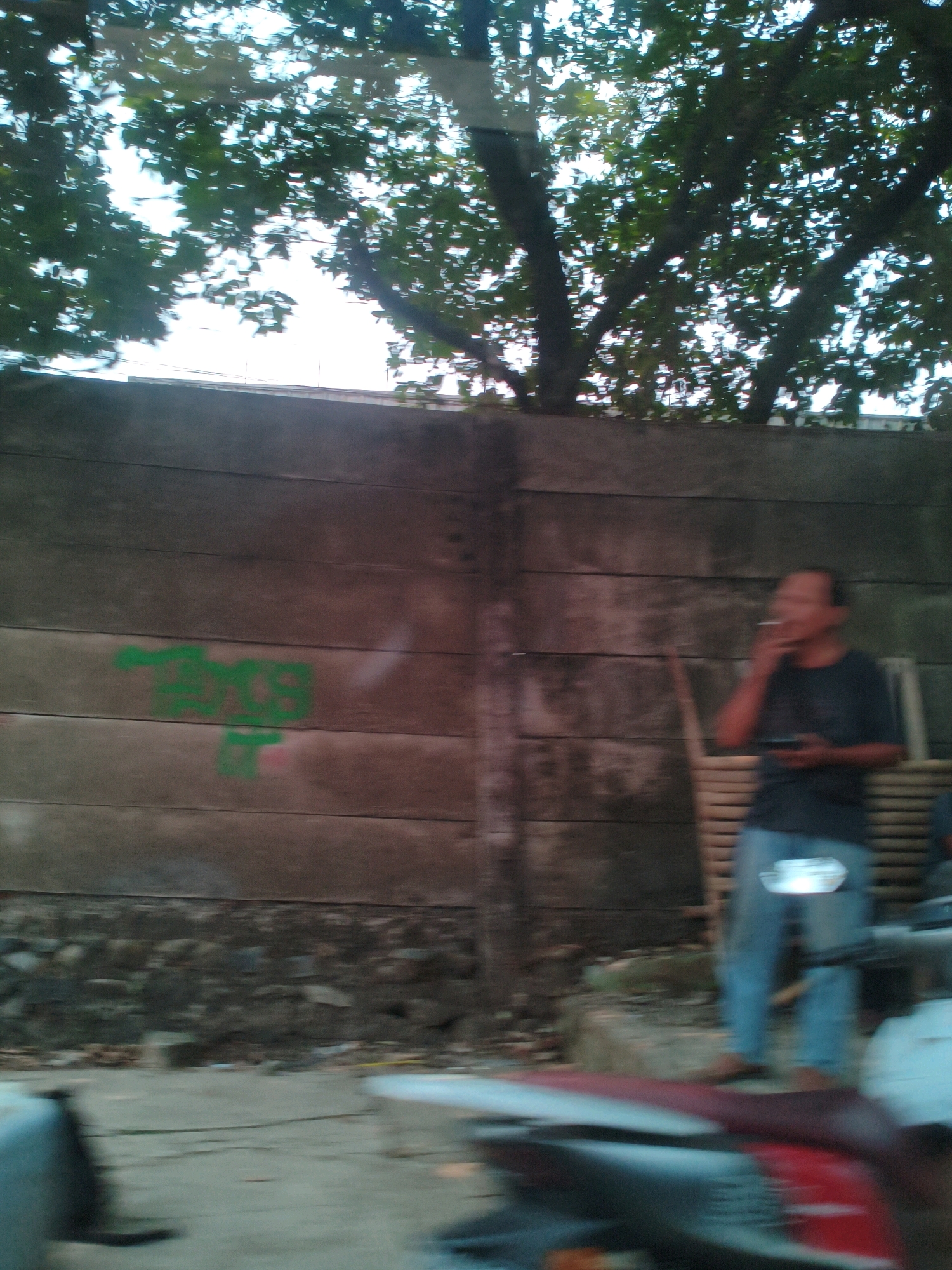 A man is standing near a concrete wall and smoking a cigarette while holding a phone. Green graffiti is visible on the wall. A motorcycle is in the foreground, and trees and foliage are in the background. The scene appears to be in Sukaraja, Indonesia.