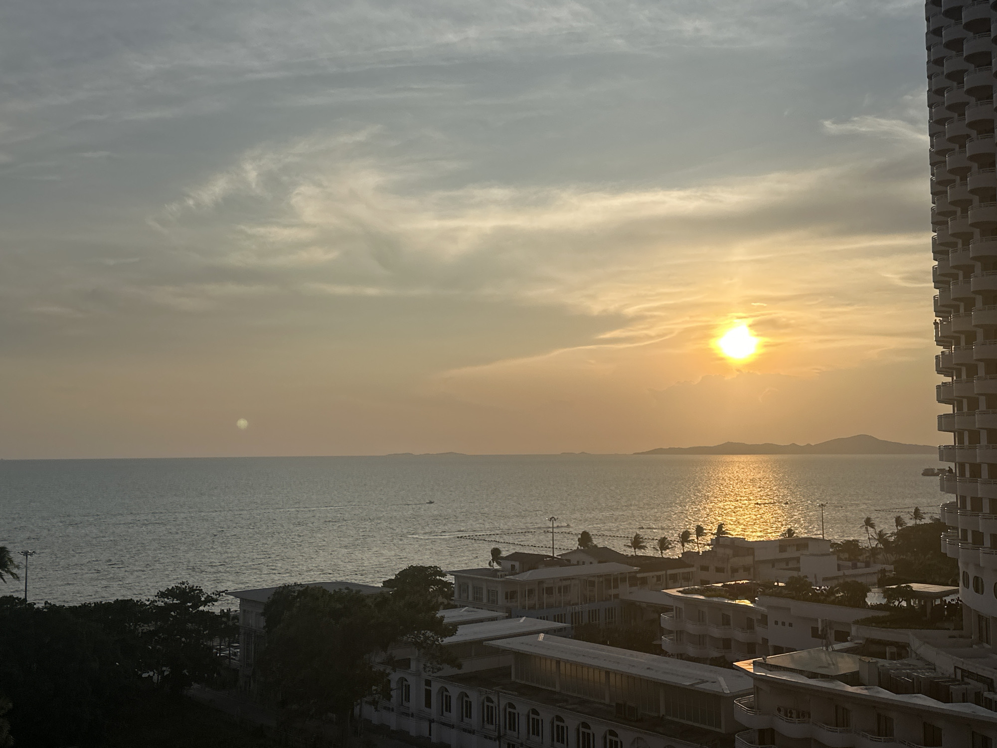 The image captures a sunset over the ocean in Pattaya City, Thailand. The sun is a bright orb, reflecting on the water's surface. Buildings are visible in the foreground, along with some trees. A tall building is located on the right. The sky has varied shades of gray and orange, with some cloud cover. The sea stretches towards the horizon, with some distant land masses. A few boats can be observed on the water.