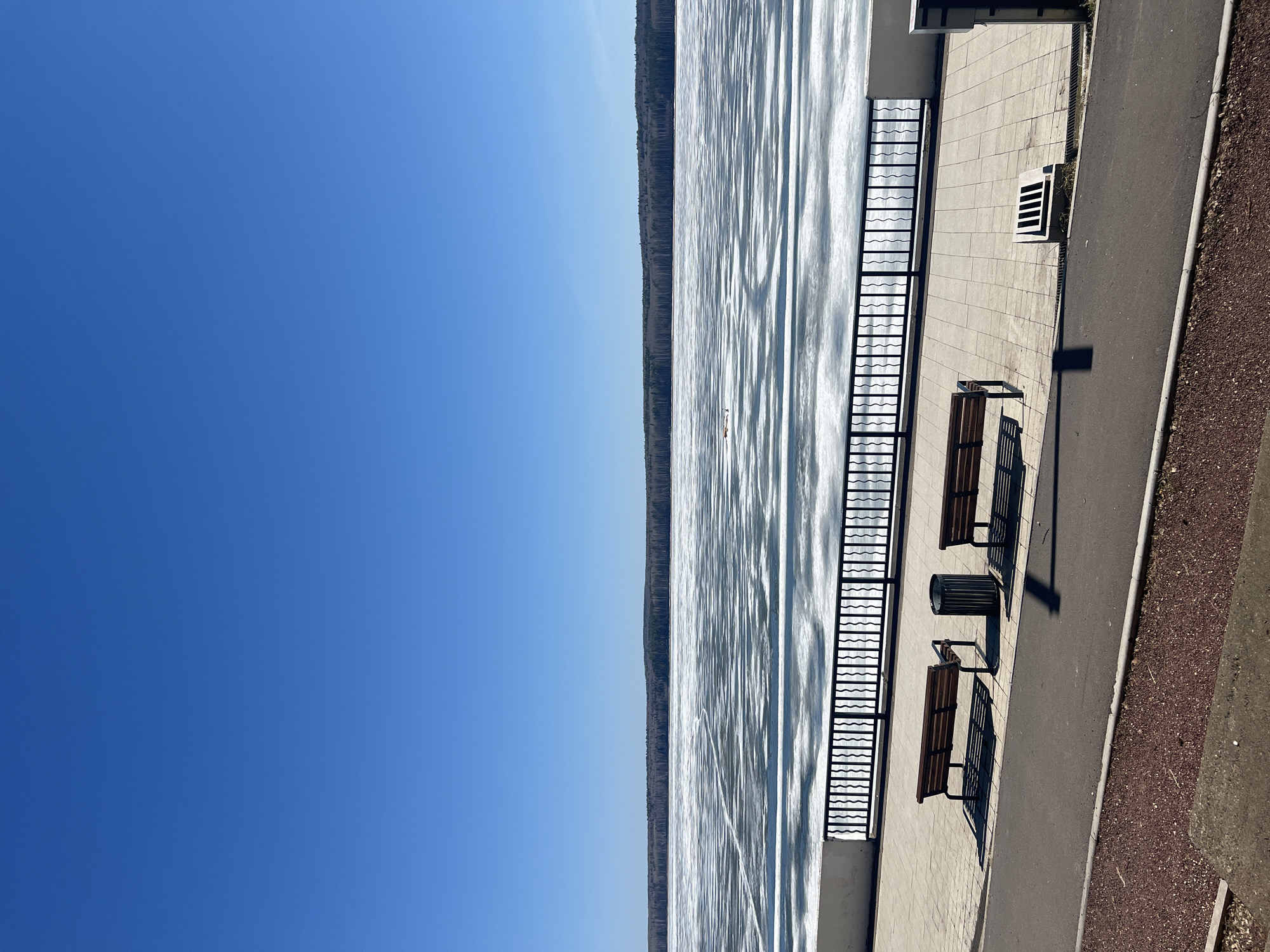 The image shows a waterfront scene on a clear day. The water is expansive and reflects the sunlight, creating a shimmering effect. Along the edge of the water is a paved walkway with a metal railing. Benches are situated along the walkway, offering seating with a view of the water. There's also a trash receptacle near the benches. Beyond the water, a line of trees can be seen along the far shore. The sky is a solid blue, with no clouds visible.