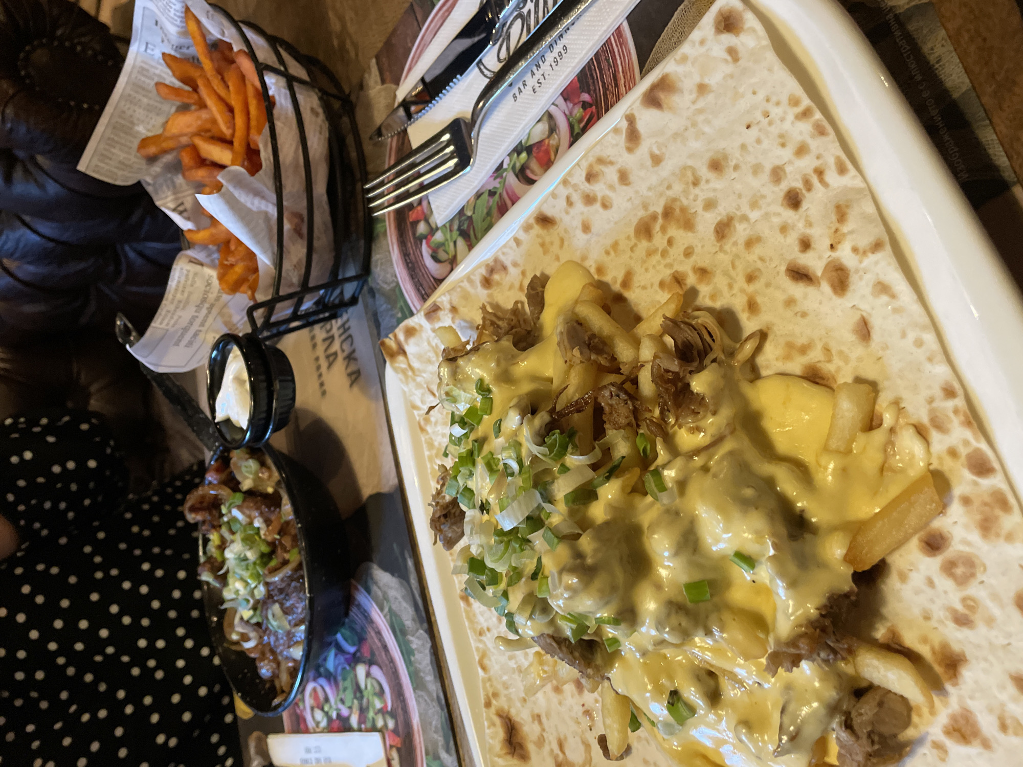 The image shows a table setting with various food items. A black wire basket contains sweet potato fries served on newspaper-patterned liners. Next to it is a small black bowl with meat, onions, and green onions, topped with white sauce. A fork and knife rest on a coaster. A large flatbread is topped with fries, meat, cheese sauce, and green onions.
The text "BAR AND DINN EST. 1999" and "ЕНСКА PAA" suggest the location may be in Veliko Tarnovo, Bulgaria.