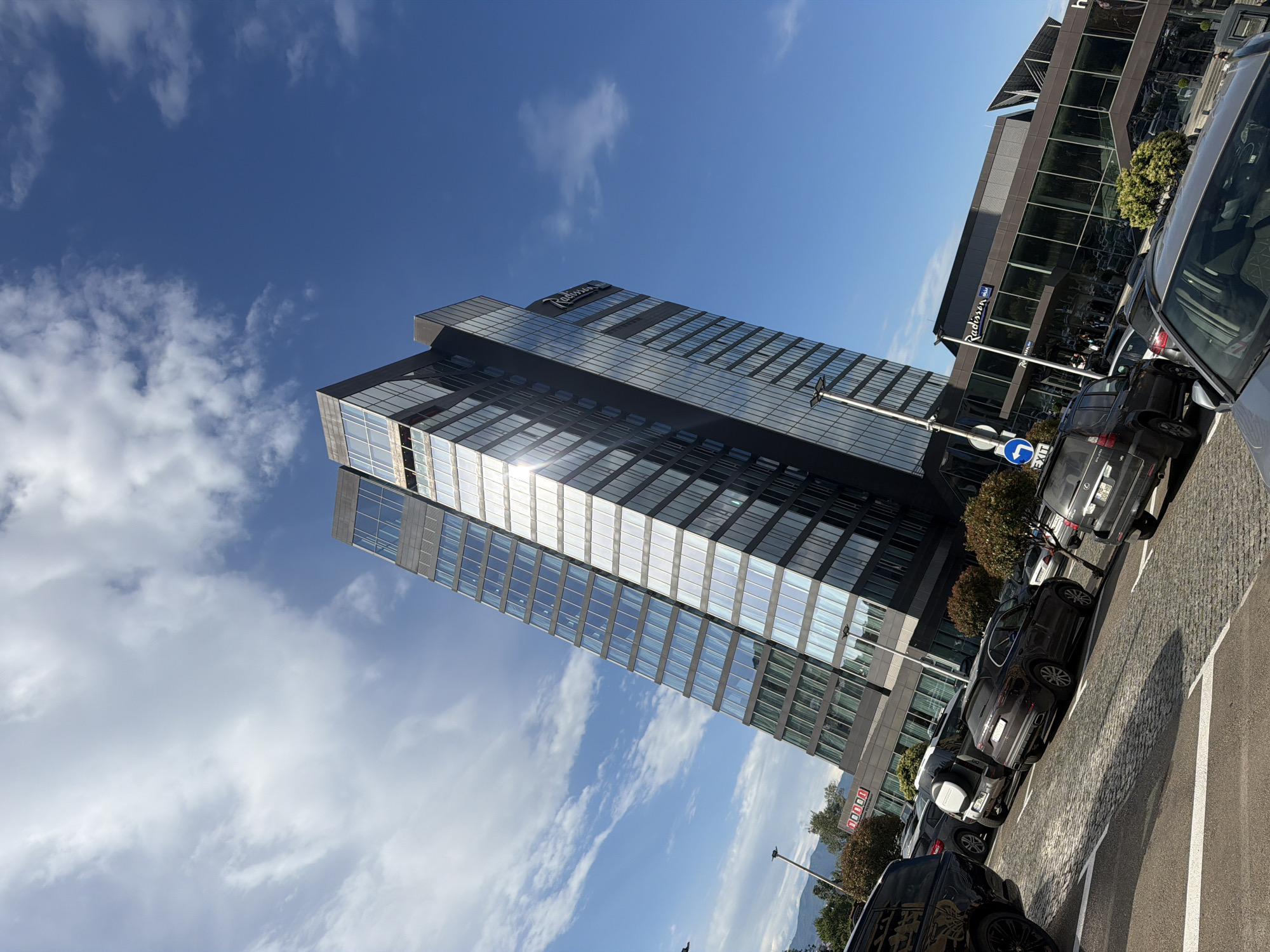 The image captures a modern urban landscape under a bright blue sky dotted with scattered clouds. The focal point is a tall, glass-clad building with a sign indicating it is a Radisson hotel. The building reflects the sunlight.
Adjacent to the hotel is a shorter, rectangular building with dark windows. The foreground features a parking area with several cars parked along white lane markings on a cobbled surface. A street sign is visible near the base of the taller building, suggesting a street intersection. The environment suggests a contemporary, possibly business-oriented district within the city.