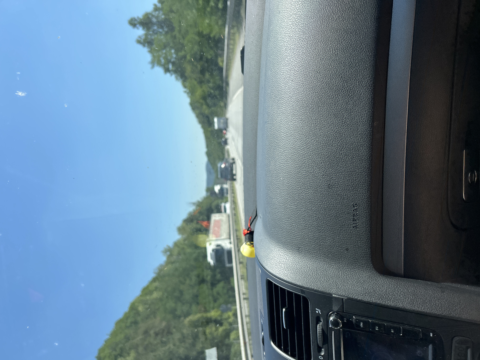 The image shows a view from inside a car driving on a highway. The dashboard of the car is in the foreground, with a textured gray surface. A yellow object with red strings is attached to the dashboard. Beyond the dashboard, the highway stretches out, with other vehicles visible. Trees and hills line the sides of the road. The sky is blue and clear. The location of Dolna Vasilitsa, Bulgaria is not discernible from the image.