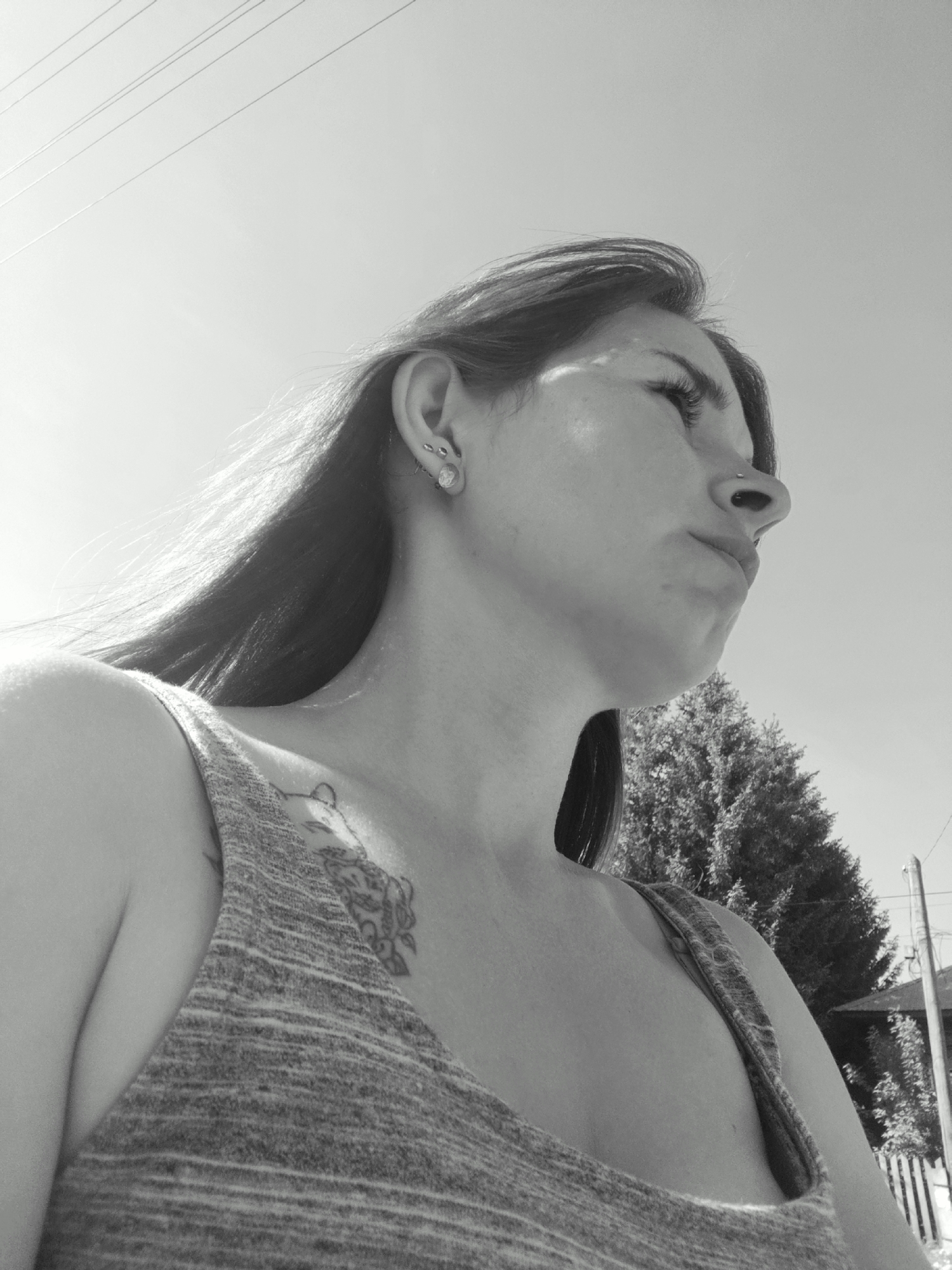A low-angle, black-and-white photograph shows a woman with long hair, facing right and upwards. She wears a tank top, and a tattoo is visible on her upper chest. She has multiple piercings in her left ear. The sky is visible above her, with power lines in the upper left corner. Trees and a portion of a building with a white picket fence are in the background to the right of the woman. The location appears to be outdoors and possibly urban. The city of Фоки, Russia, cannot be verified.