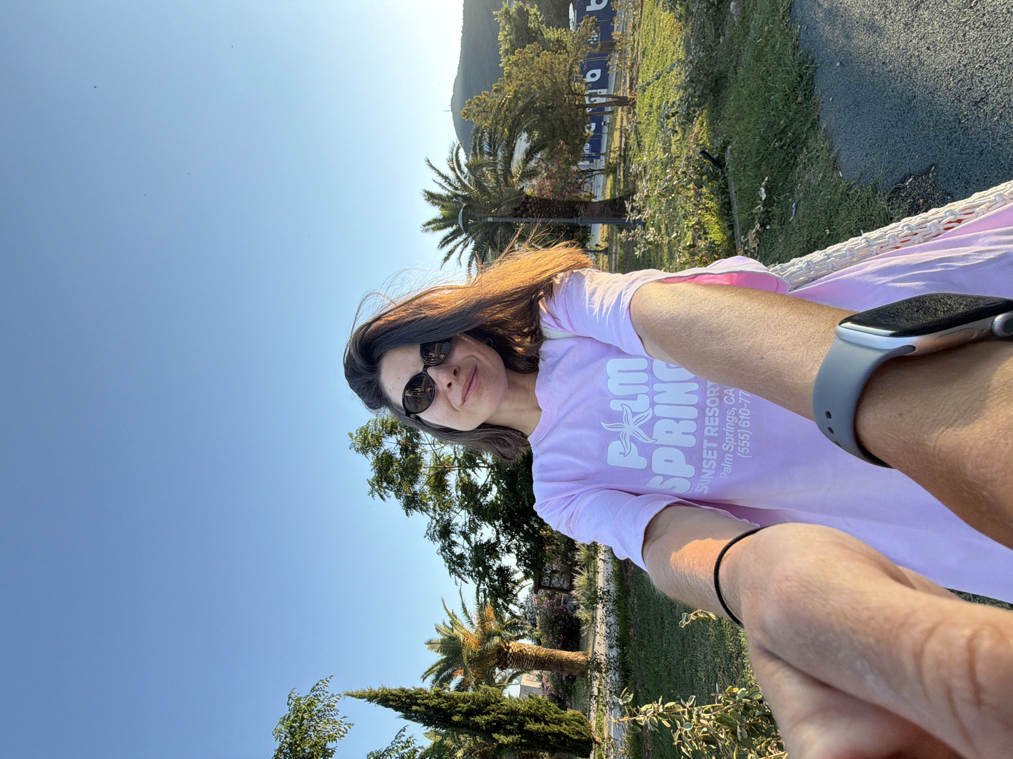 The media file is a selfie taken outdoors on a sunny day, likely in the late afternoon given the warm light. The subject is a woman wearing sunglasses and a light purple t-shirt with white text that reads "SUNSET RESORT Palm Springs, CA (555) 610-77". She is smiling and appears relaxed. Her right arm, wearing a grey smartwatch, is extended towards the camera. A black bracelet is visible on her left wrist.
The background shows a lush green environment with several palm trees and other foliage, suggesting a warm climate. In the distance, a hillside covered in trees can be seen. The setting appears to be a park or resort area with a paved path and grassy areas. The sky is a clear, bright blue. The orientation of the image suggests it was taken while the person was lying down or seated, perhaps in a hammock or lounge chair, given the angle of the arm and the visible white woven material in the foreground. The overall mood is one of leisure and enjoyment of a pleasant day outdoors. The location context of Budva, Montenegro, aligns with the visual cues of sunshine, palm trees, and a Mediterranean-like landscape.