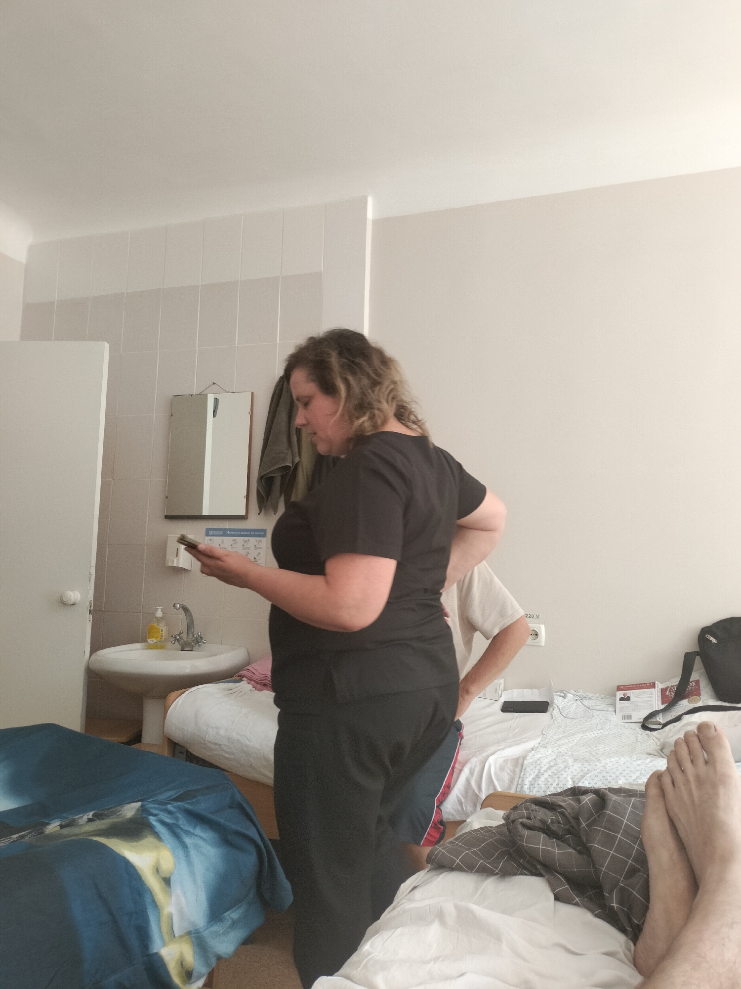 The image depicts an interior scene, likely a hospital room in Chernihiv, Ukraine. A woman with curly brown hair is standing in the foreground, wearing a black t-shirt and black pants. She is holding a smartphone and looking down at it. Behind her, a portion of another person wearing a light-colored t-shirt and red-striped shorts is visible. The room contains two beds, one with blue bedding featuring a landscape print, and another with white bedding. A sink with a faucet and a yellow soap dispenser is visible on the left. A mirror hangs above the sink, reflecting a gray towel. A white door is to the far left. The walls are tiled in a light color, and a wall socket labeled "220 V" is present. A black backpack and some books are on the bed in the background. The feet of a person are visible in the bottom right corner of the image, lying on a bed with grey and white checkered blankets.