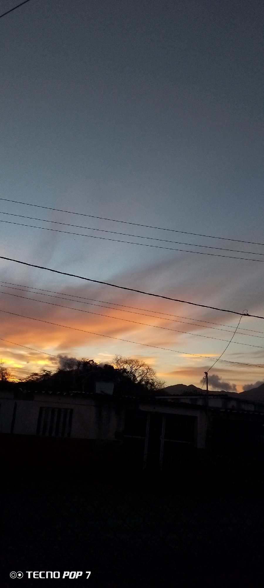 A vertical shot depicting a sunset scene in Altagracia, Venezuela. The sky shows hues of orange, yellow, and blue, partially obscured by power lines. In the foreground, a dark silhouette reveals a structure, possibly a building, with a tree line and faint mountain shapes behind it. The lower edge of the image is mostly black. The watermark "TECNO POP 7" is visible in the lower-left corner.