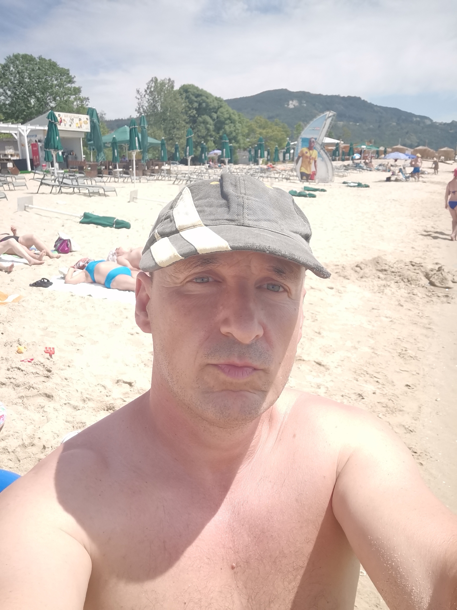 The image depicts a man wearing a grey cap and no shirt, posing for a selfie on a sandy beach in Kranevo, Bulgaria. In the background, beach chairs, umbrellas, and a small beach bar are visible. The sea is not visible in the frame, but the background suggests a mountainous landscape. Several other beachgoers are present, some lying on chairs or towels, and others walking on the sand. The sky is partly cloudy.