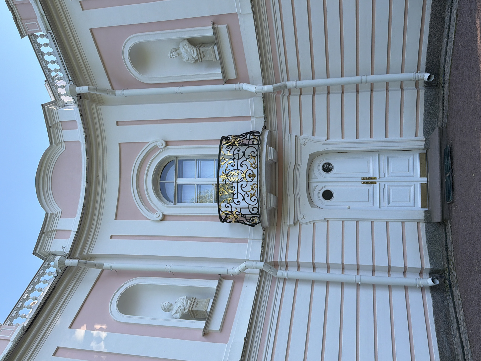 The image shows a pink and white building with an ornate balcony. The building has a curved facade, with a central arched window above a pair of double doors. The balcony has a black metal railing with gold decorative elements. On either side of the balcony, there are niches with white busts of people. The building is surrounded by a gravel area, and the sky is visible in the background. The architectural style appears to be Baroque or Rococo.