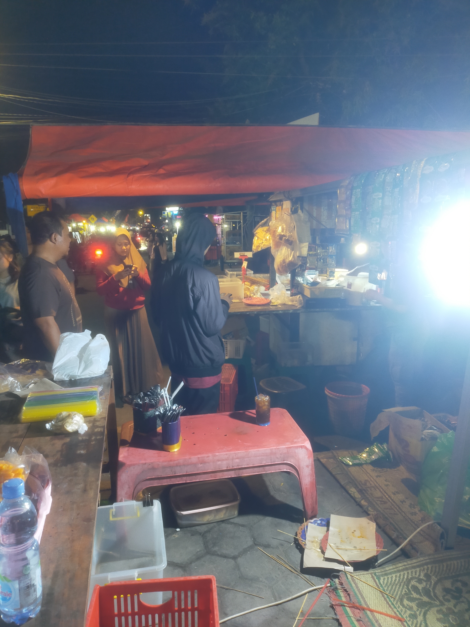 In Bantul, Indonesia, at night, an outdoor food stall operates under a large orange tarp. Three people are visible. A man in a dark t-shirt stands on the left, facing right, with his back mostly to the camera. A woman wearing a hijab and a reddish-pink long-sleeved top and a brown skirt stands in the center, looking at her phone. A person wearing a dark hooded jacket and dark pants stands on the right, facing the stall. The stall features a table with various items including a white plastic bag, colorful straws, and snacks in plastic packaging. A red plastic table sits in front of the stall with a glass of dark liquid and a container of utensils. Behind the table, a shelf is laden with bottles, food items in plastic bags, and other merchandise. The ground is covered with mats. The scene is illuminated by bright lights, possibly from the stall and surrounding streetlights, creating a well-lit but somewhat hazy atmosphere.