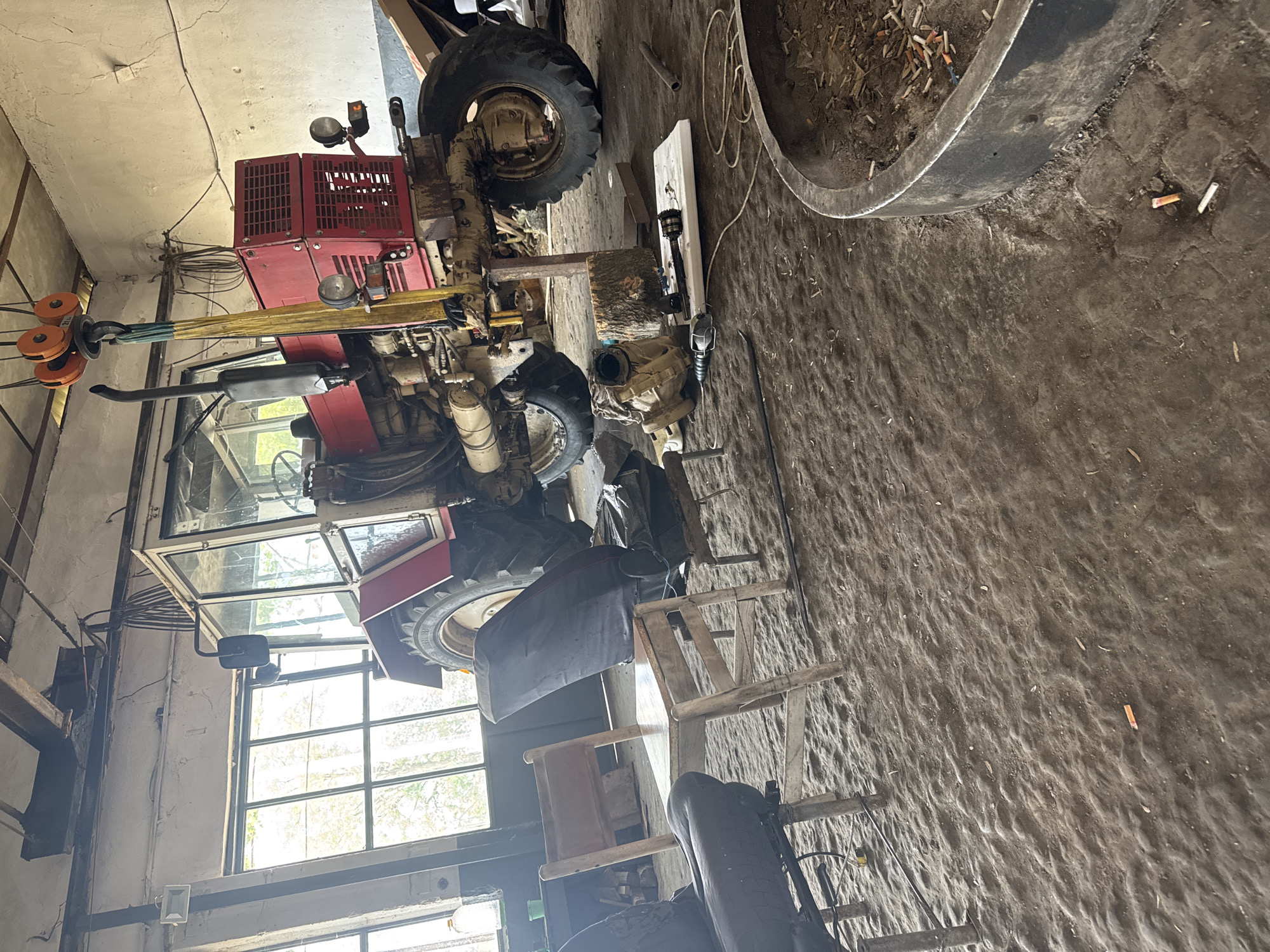 The image captures an indoor scene, likely a rural workshop, garage, or barn in Sarnevo, Bulgaria, during the daytime. The primary subject is a large, red tractor, possibly a Belarus or similar Eastern European model, which appears to be undergoing maintenance or repair.
The tractor is prominently featured, positioned towards the left of the frame. Its engine compartment and front-right side are visible, with a large, lugged tire on the front axle and another, partially visible, rear tire. An exhaust pipe extends upwards near the cabin. A yellow strap and orange pulleys of a lifting device or hoist are attached to the front section of the tractor, suggesting a component is being lifted or supported.
The interior of the space has light-colored walls and ceiling, showing some cracks and wear. A large window with multiple panes, located behind the tractor's cabin, allows natural light to illuminate the scene. The floor is made of concrete, appears textured and uneven, and is covered with dirt and miscellaneous debris.
Several other objects are scattered around the workshop. On the right, a large, dark, circular metal rim, possibly from a large tire or an industrial container, is filled with dirt and numerous cigarette butts. More cigarette butts are visible scattered on the concrete floor. A wooden ladder leans against a surface near the tractor's rear. Other mechanical parts, including a driveshaft and what looks like a transmission or axle component, are resting on the floor or on a wooden block next to the tractor, further indicating ongoing work. Cables and electrical wiring run along the walls and ceiling.
No people are visible in the image. There is no discernible text. The bright lighting through the window suggests it is daytime. The overall impression is that of a functional, utilitarian workspace for agricultural machinery.