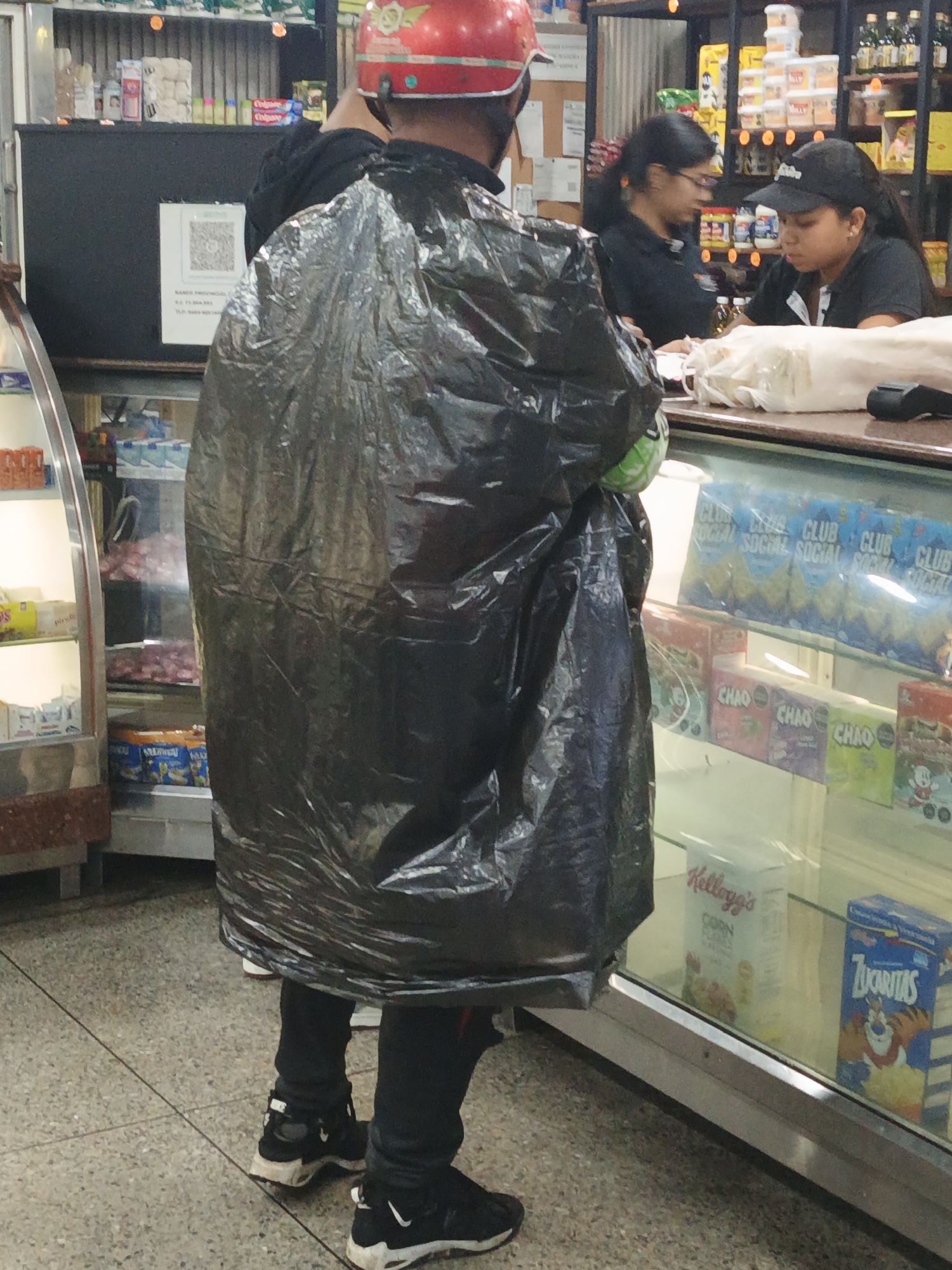 A person wearing a red motorcycle helmet and covered in a large black plastic bag stands facing a store counter. Two women stand behind the counter; one with glasses and another wearing a black baseball cap. The counter has a display case containing snacks and cereal, including Club Social crackers, Chao candy, Kellogg’s Corn Flakes, and Zucaritas cereal. Behind the counter are shelves stocked with various goods. The scene is located in Tinaquillo, Venezuela.