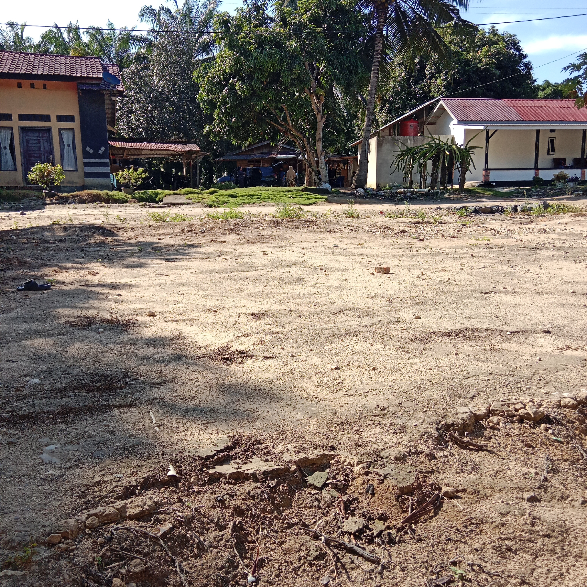 The image depicts an outdoor scene in a location that appears to be a rural or semi-rural area, likely in Indonesia given the vegetation and architecture. In the foreground, there is a dirt and gravel ground with patches of dry earth and scattered debris, including a single dark-colored sandal on the left side. The ground shows signs of unevenness and erosion in places.
In the middle ground and background, there are several buildings and dense vegetation. On the left, a two-story house with a beige and dark-colored facade, a red tiled roof, and windows is visible. Adjacent to it is a covered outdoor area or a small building with a corrugated roof. Further to the right, a larger, single-story building with a white facade and a red corrugated roof is prominent. This building has a covered porch supported by pillars. There is a red water tank visible on top of a part of this building. A cluster of dragon fruit plants is growing in front of this building. Between the buildings, there are trees with lush green foliage, including palm trees, suggesting a tropical climate. In the area behind the buildings, more trees and vegetation are visible, forming a dense backdrop.
Several individuals are present, mostly clustered around an area with a covered shelter and a vehicle. Some appear to be standing or walking. The overall impression is a residential area with a mix of simple housing and natural surroundings. The lighting suggests it is daytime, with sunlight casting shadows across the ground.