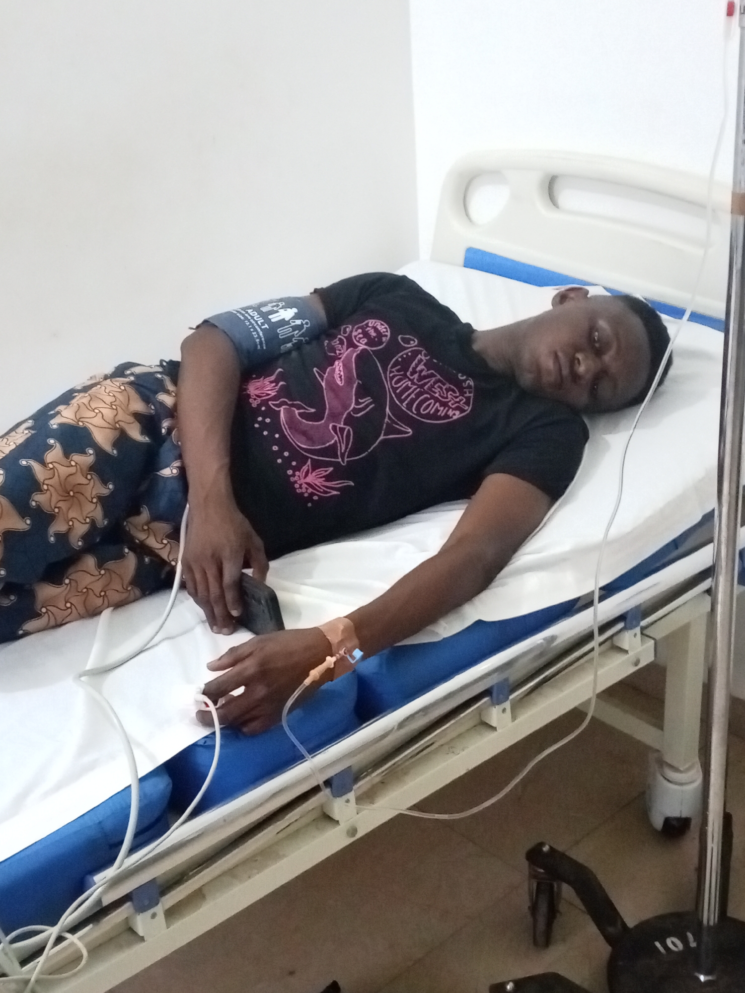 A young man lies in a hospital bed with an IV drip connected to his left arm. He wears a black t-shirt with a graphic design and patterned trousers. A blood pressure cuff is on his upper left arm, and he holds a small device in his left hand. The bed has white sheets with a blue border. An IV pole stands beside the bed. The location is potentially a medical facility in Sabon Tasha, Nigeria.