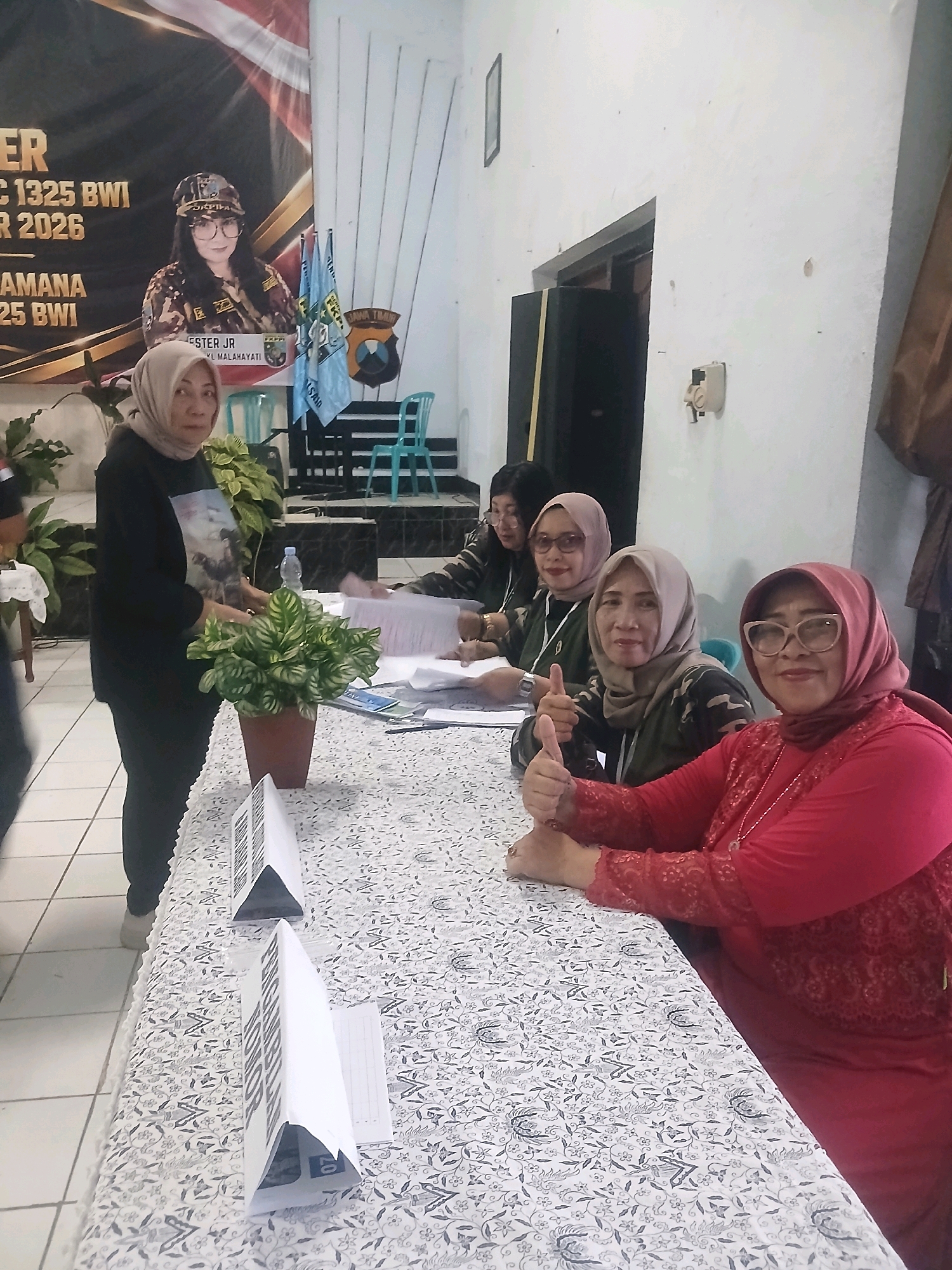 In a room that appears to be an indoor community hall or office space in Banyuwangi, Indonesia, several women are gathered around a table draped with a patterned cloth. The room has white walls and a tiled floor. A large banner is visible in the background, featuring text and a portrait of a woman in a cap and camouflage attire. The overall scene suggests an event or a registration desk, possibly related to a local organization given the presence of flags and organizational imagery.
In the foreground, a woman wearing a red hijab and a matching red long-sleeved top with lace detailing sits at the table. She is smiling and giving a thumbs-up gesture. Beside her, another woman in a light brown hijab and a camouflage jacket also offers a thumbs-up. To her left, a third woman in a darker brown hijab and a similar camouflage jacket is also seated, looking towards the camera.
A woman in a black hijab and black outfit stands to the left of the table, looking towards the camera. She is holding a potted plant with green and yellow variegated leaves. In front of her, a water bottle and various papers are arranged on the table. Triangular signs with black text on a white background are placed on the table as well, suggesting directional markers or labels.
In the middle ground, several chairs, possibly plastic and teal in color, are visible, along with what appears to be a stage or raised platform. Two flags are displayed; one is blue with white writing and a logo, and the other is a red and white flag, likely the Indonesian flag, though its details are obscured. The environment is functional, with basic furnishings and decorations, indicative of a local meeting or administrative setting.