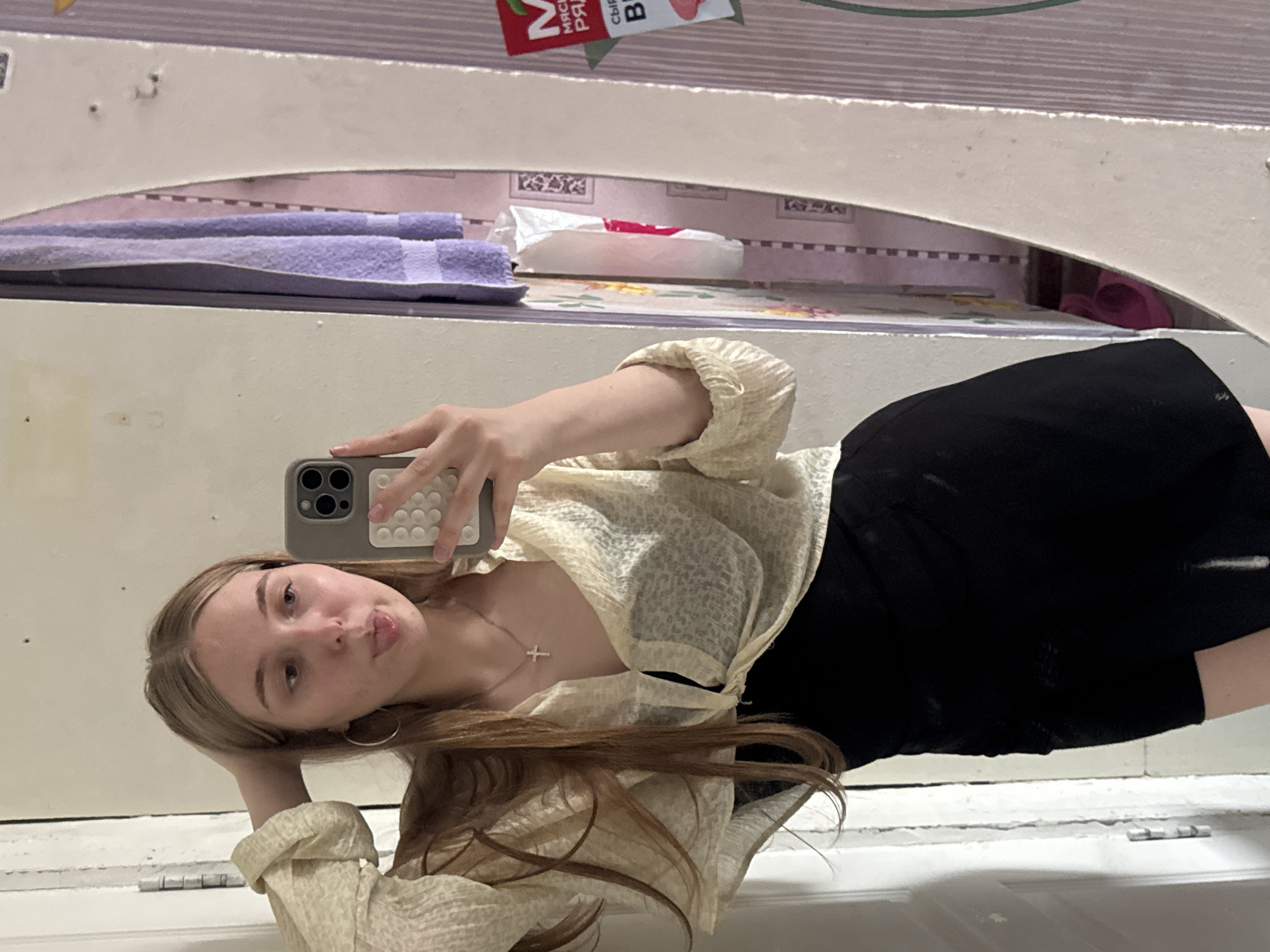 A young woman with blonde hair is taking a selfie in a dimly lit room. She is wearing a sheer, cream-colored blouse tied at the waist over a black dress. A silver cross necklace is visible. Her left hand is raised to her head, and her tongue is sticking out playfully. She is holding a smartphone in her right hand. The background shows a door on the left and a mirror on the right. In the mirror's reflection, two purple towels are hanging, and a colorful piece of wallpaper or fabric with a checkered pattern is visible. A red and white package is also reflected in the mirror. The room appears to be a domestic setting. Based on the context of the reflection of a package with Russian text and the cultural context suggested by the image style, this scene is likely located in Perm, Russia.