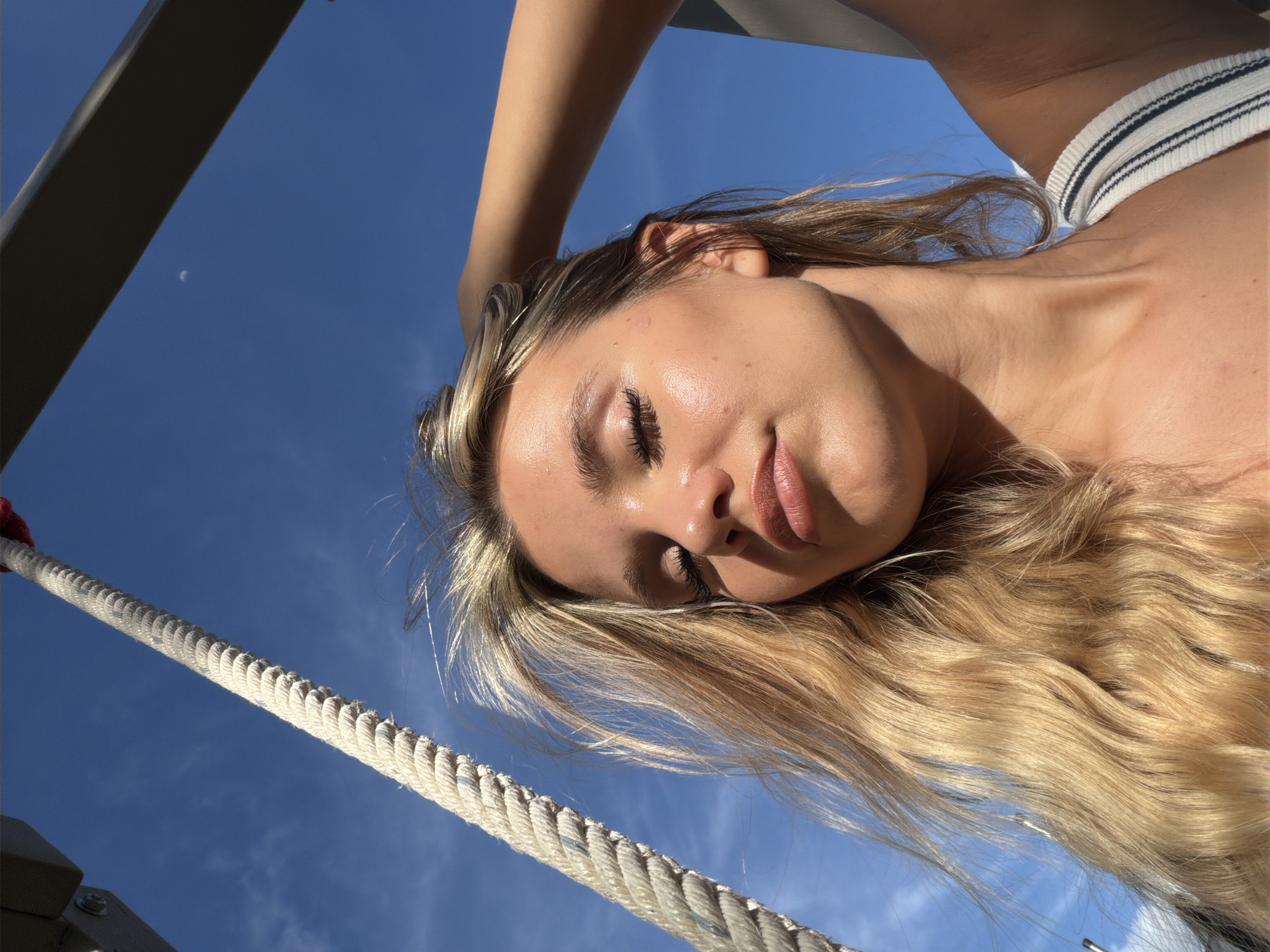A young woman with long, blonde hair takes a selfie outdoors against a bright blue sky. She is positioned in the lower right quadrant of the frame, with her head tilted upwards and her eyes closed, one hand raised to adjust her hair. A thick, white rope runs diagonally across the left side of the image, and a dark, angular structure is visible at the top right. The sunlight casts highlights on her skin. The background shows a clear sky with a faint crescent moon visible. This scene appears to be captured in the city of Thep Krasatti, Thailand.