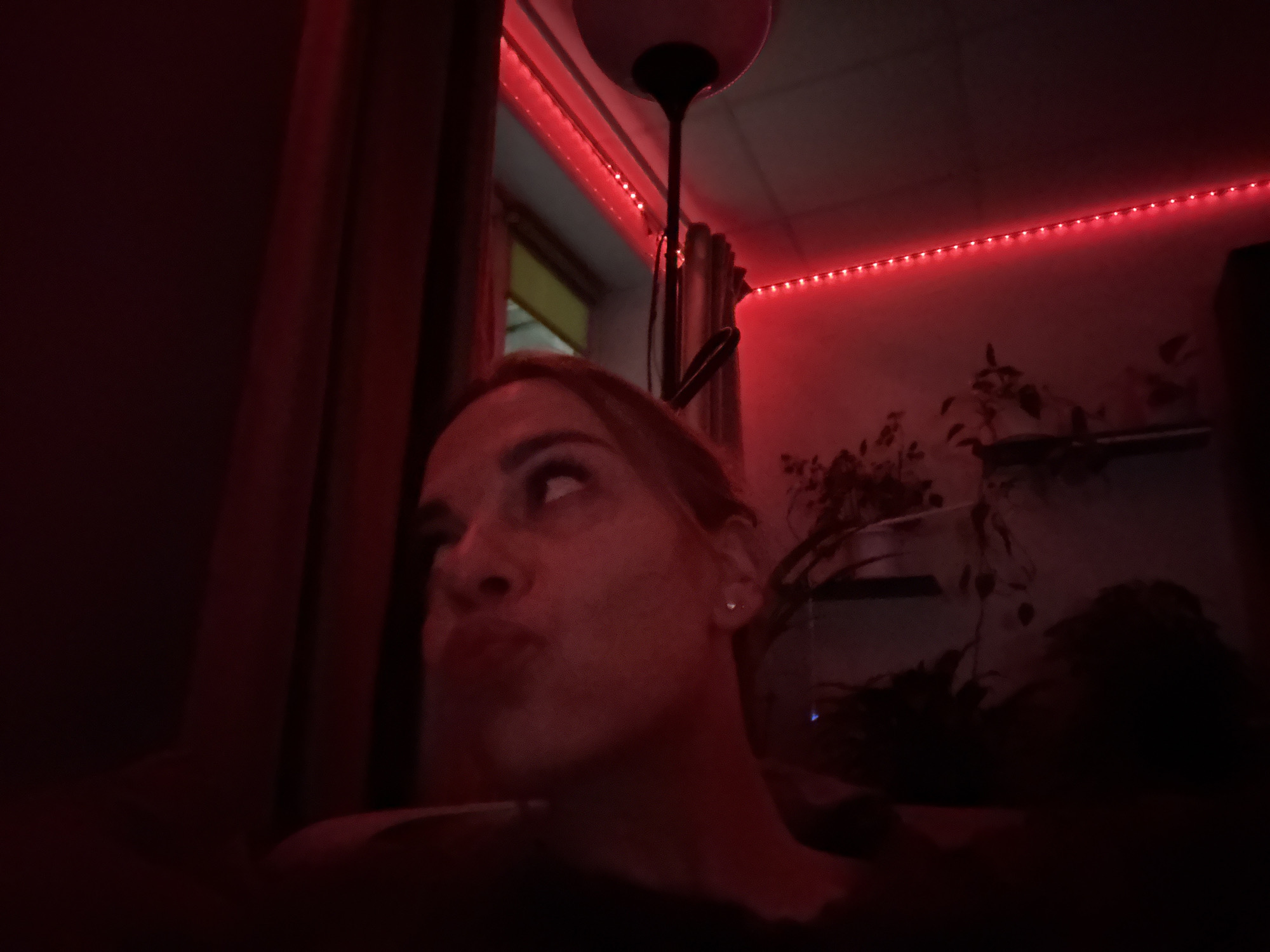 A low-angle shot shows a person with light skin and light-colored hair, illuminated by red lighting. The person's face is turned slightly upwards, and their lips are pursed. Above the person, red LED strip lights run along the ceiling and window frame. A floor lamp is also visible. In the background, there are plants. The photo likely originates from an indoor setting. It is not possible to determine the exact location of Ust-Ilimsk, Russia from this image.