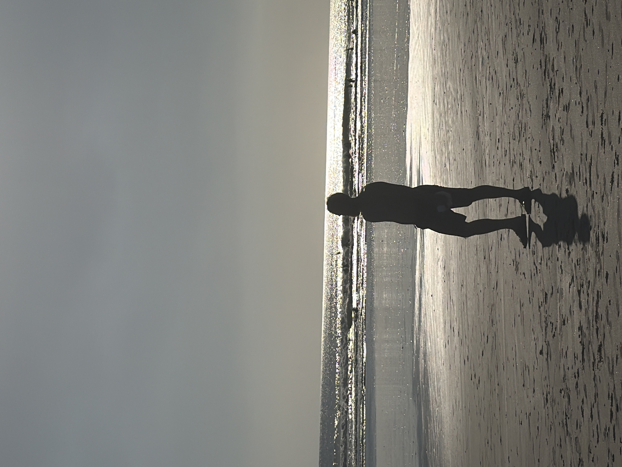 A silhouette of a man stands on a sandy beach, facing the ocean. The sun is low in the sky, casting a bright glare on the water and creating a reflection on the wet sand. The man is wearing shorts and appears to be looking out at the waves. In the distance, several people can be seen in the water, some appearing to be swimming or surfing. The sky is clear and overcast, with a light haze. The scene suggests a tranquil, late afternoon at the beach, possibly Kuta, Indonesia, known for its surfing beaches and vibrant atmosphere.