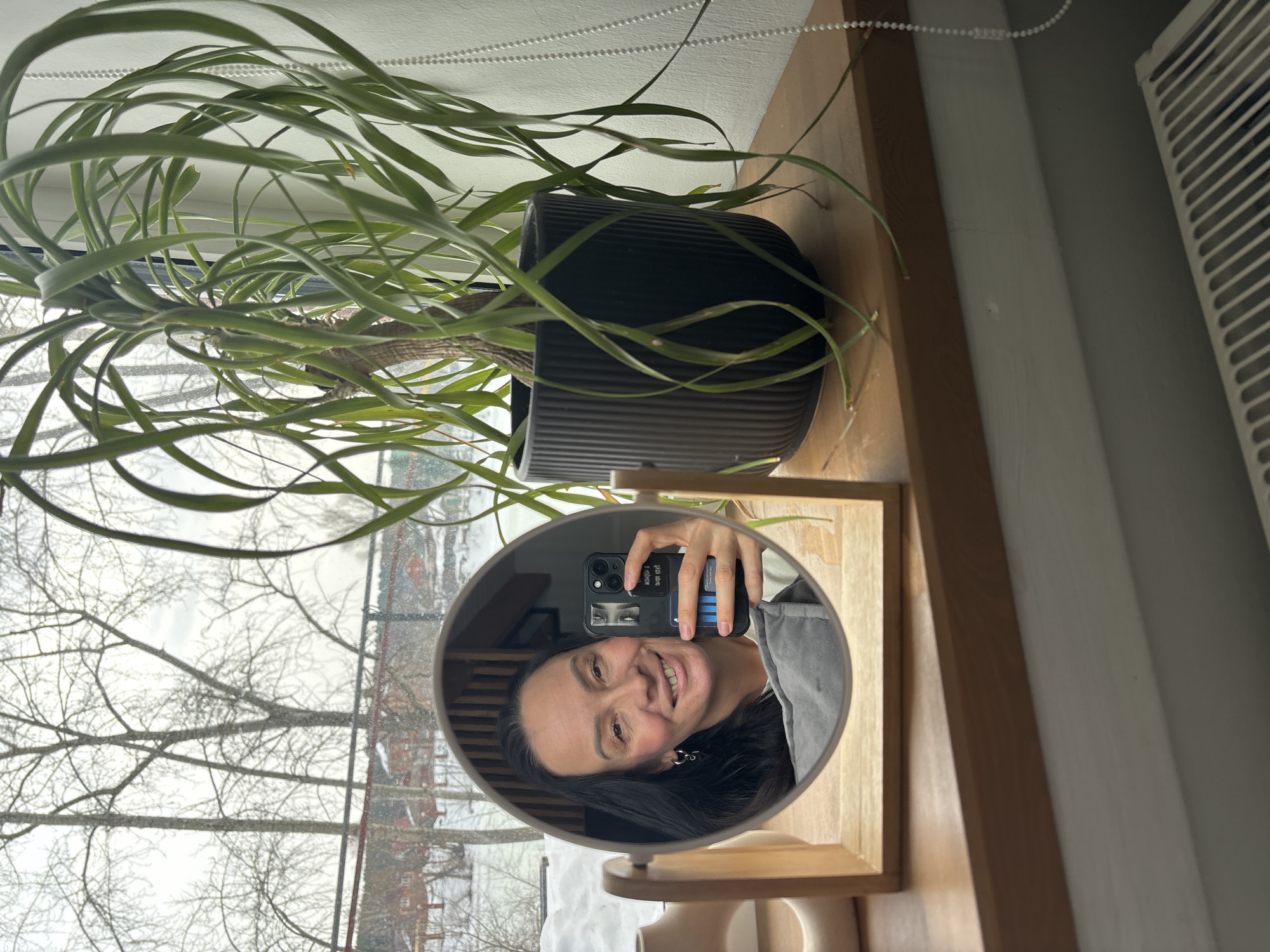 An indoor scene in Chaikovsky, Russia, features a selfie taken in a circular mirror on a wooden windowsill. The mirror reflects a woman with long, dark hair, smiling and holding a black smartphone with a distinctive case. A blue card is visible behind the phone. She wears a light-colored top and earrings. A gray pillow or blanket is partially visible below her reflection. To the right of the mirror, a large potted plant with long, drooping green leaves sits in a dark, ribbed cylindrical pot. The windowsill also holds a light-colored decorative object to the left of the mirror. Through the window, a winter landscape is visible, characterized by snow-covered ground and numerous bare deciduous trees. Distant red-roofed buildings are interspersed among the trees, and a dark horizontal structure, possibly a fence or railing, spans the mid-ground. The sky is overcast. A white beaded chain for blinds hangs on the right side of the window, and a white radiator is partially visible below the windowsill.
