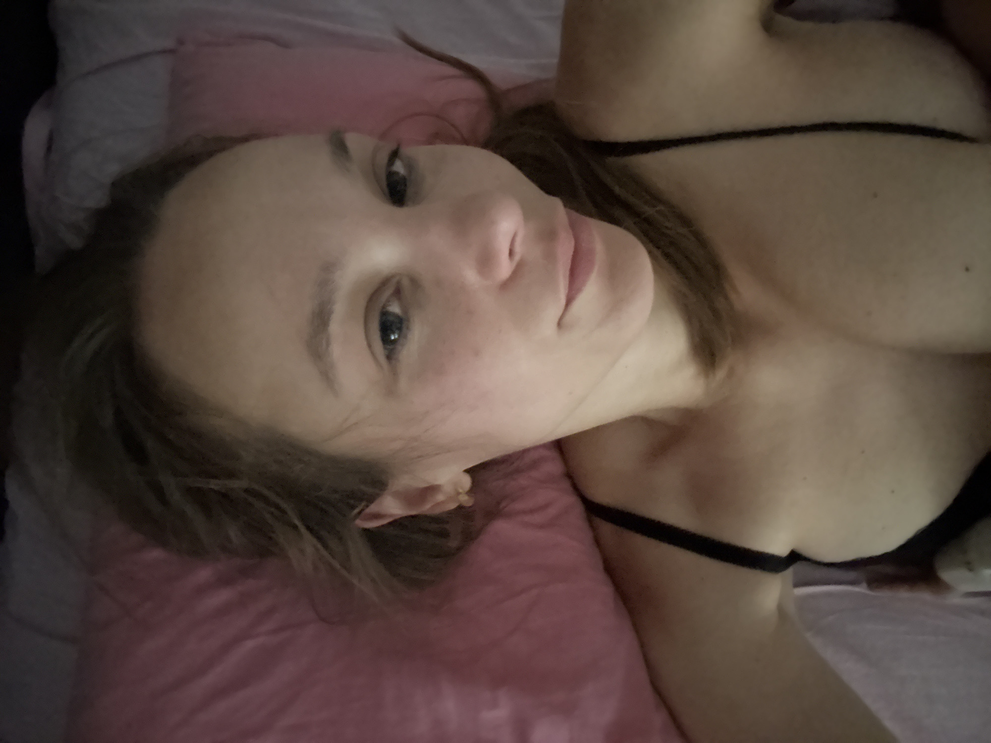 The image is a close-up, selfie-style photograph of a young woman lying down, appearing to be in a bed with a pink pillow. She is looking directly at the camera with a relaxed expression. Her hair is down, partially obscuring her face and shoulders. She is wearing a black bra with thin straps. The background is softly blurred, suggesting an indoor setting. There is no discernible information within the image that would indicate a specific location in Überherrn, Germany.