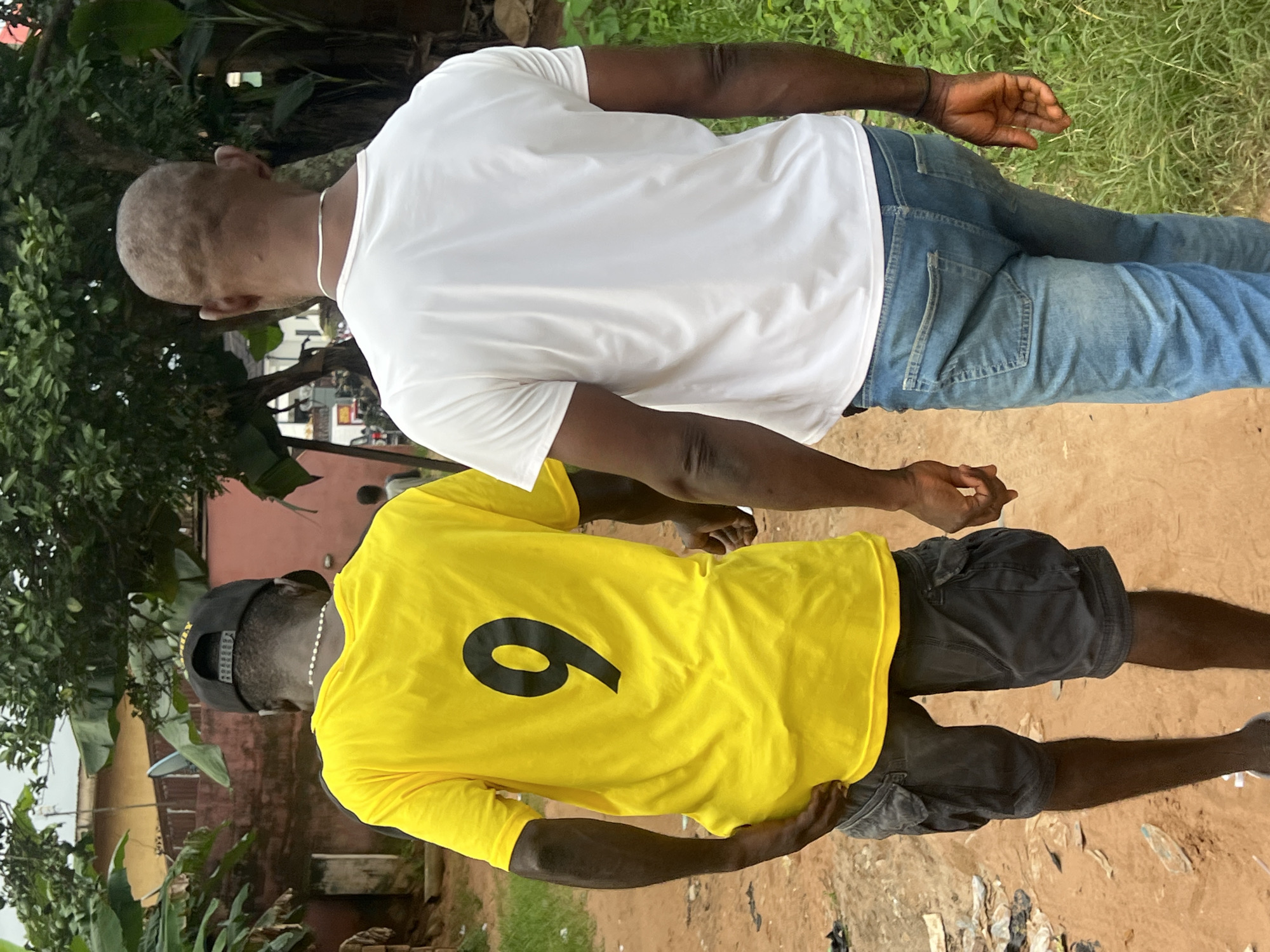 In Nkpor, Nigeria, two men walk away from the camera on a dirt path. The man on the left wears a bright yellow t-shirt with a large black number "9" on the back, a black baseball cap, and dark cargo shorts. The man on the right wears a plain white t-shirt and light blue jeans. They appear to be holding hands or walking closely together. The environment is outdoors, with green vegetation on the right side of the path and buildings and trees visible in the background. The path is unpaved and shows signs of wear and tear, with some scattered debris visible. The overall scene depicts a casual walk in a somewhat undeveloped urban or semi-urban area.
