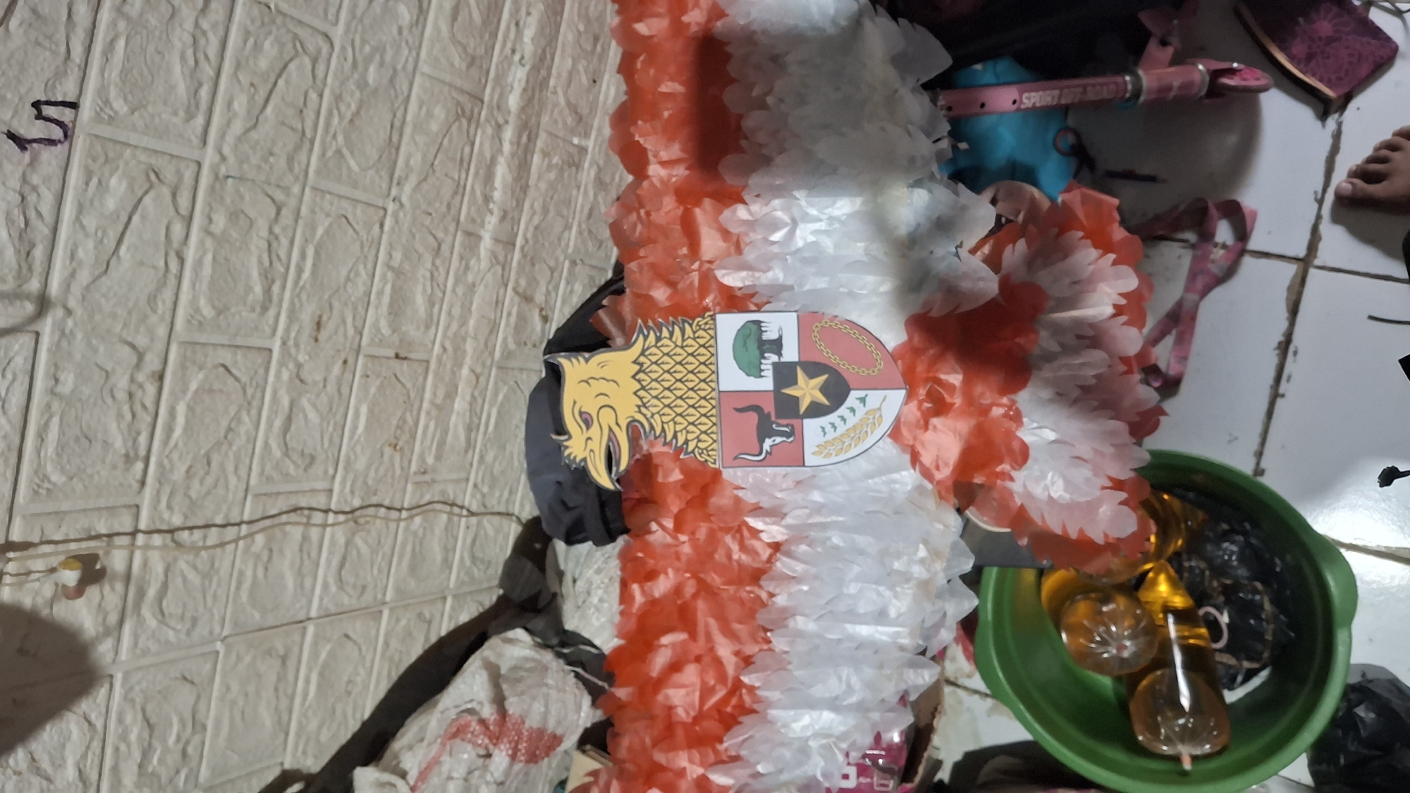 The image shows an indoor scene with a focus on a decoration resembling the Indonesian Garuda Pancasila. The decoration is made of red and white tissue paper and features the Garuda Pancasila emblem at its center. The emblem includes a yellow eagle with the shield divided into four quadrants, each containing symbols of Indonesia's national ideology.
The decoration is placed against a white brick-patterned wall and is surrounded by various objects. To the left of the decoration, there's a green plastic bowl containing several amber-colored lollipops and some black, crumpled plastic bags. Behind the decoration, out-of-focus items include what appears to be fabric or sacks, and dark-colored bags. To the right, a pink scooter with the words "SPORT..." is partially visible. The floor is tiled and appears somewhat dirty. A person's bare foot is visible in the lower right corner of the frame. The lighting suggests an indoor setting, possibly in a home or storage area.