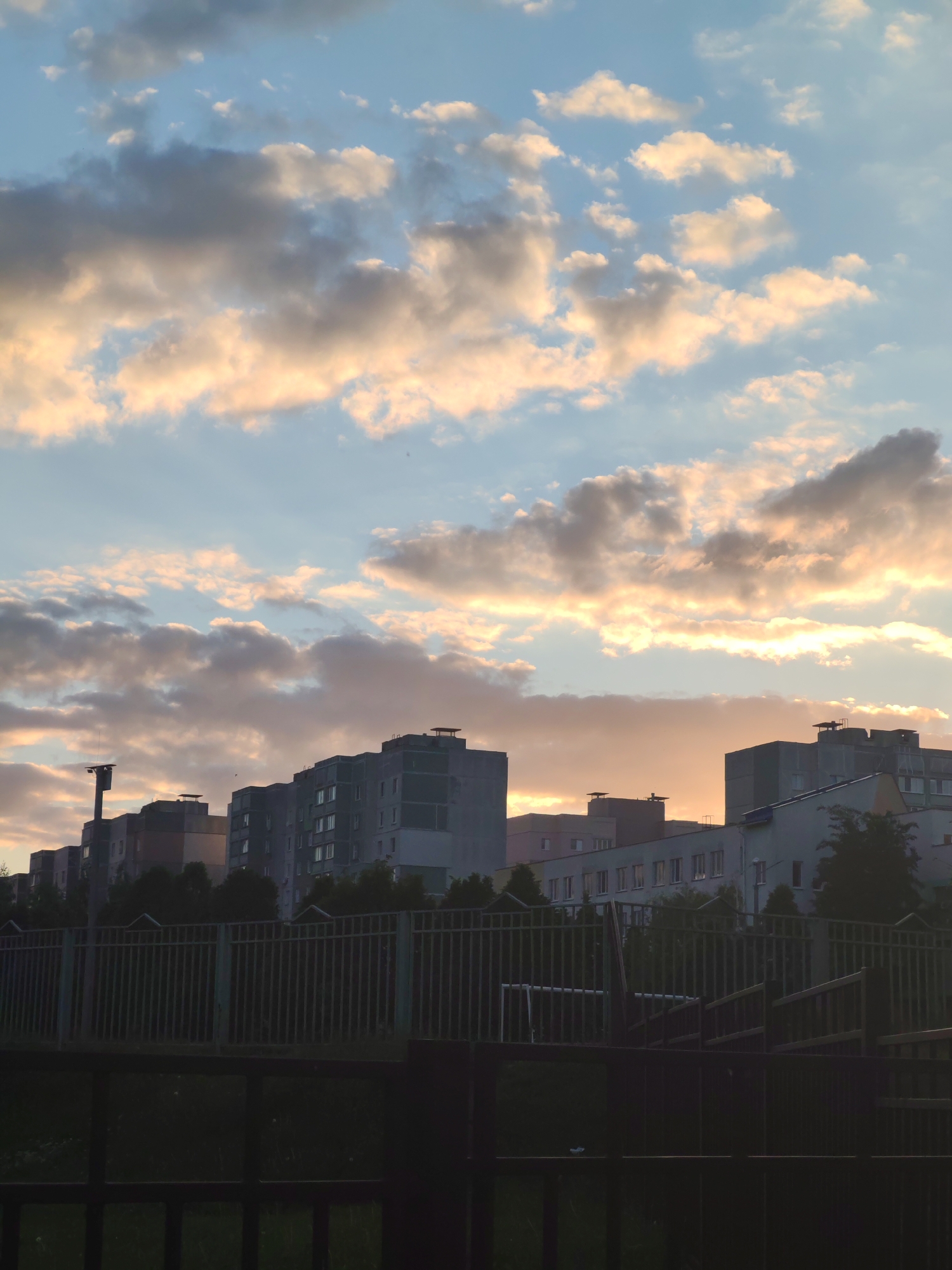 The image shows a cityscape at dusk or dawn, with a focus on the sky and buildings. The sky is filled with clouds illuminated by the setting or rising sun, creating a mix of colors including blues, grays, and oranges. Below the sky, a row of buildings is visible, appearing as silhouetted blocks against the bright sky. A fence runs along the base of the frame, obscuring a dark space with some vegetation visible through the gaps. In the middle ground, there appears to be a soccer goal partially visible behind the fence. The location appears to be an urban area with multi-story residential or commercial buildings.