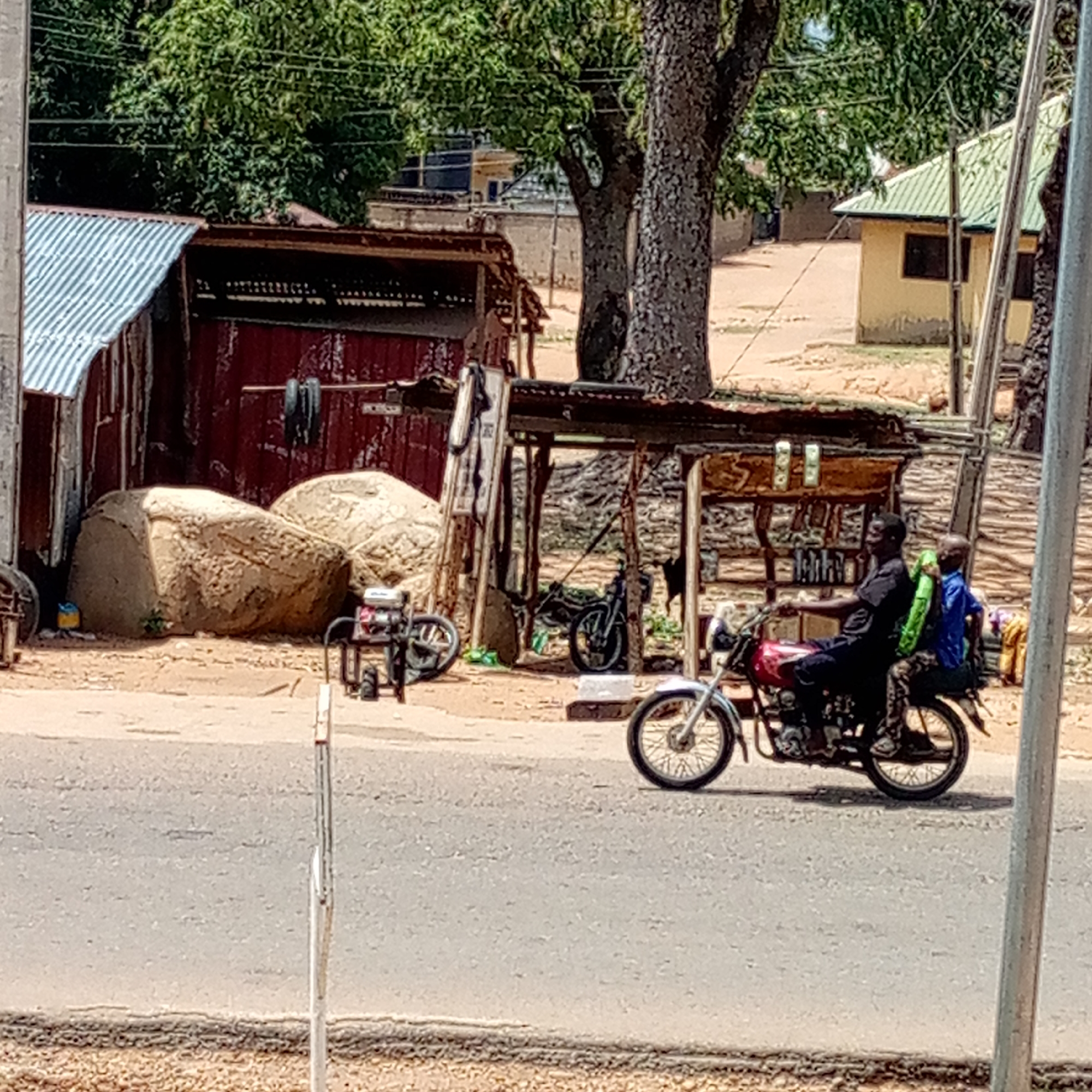 The image depicts a bright, sunny street scene in what appears to be a rural or semi-urban area. In the foreground, a man wearing a dark shirt is riding a red motorcycle, with a child seated directly behind him. The child is wearing a blue top and holding a bright green object, possibly a backpack or bundled item. They are on a paved road that shows signs of wear.
To the left of the road, the background features several structures. A building with a corrugated metal roof and reddish-brown, possibly corrugated iron, walls stands next to two large, light-colored rocks. A small, portable generator or engine unit is visible on the dusty ground near these rocks. Adjacent to this, a wooden lean-to structure with a corrugated roof provides shelter, under which another motorcycle is parked. Various items hang from or are placed within this lean-to.
Lush green trees are prominent throughout the background, indicating ample vegetation. Further in the distance, a light yellow building with a distinct green corrugated roof can be seen, partially obscured by trees and other structures. The ground immediately beside the paved road is unpaved and appears dusty or sandy. A thin metal pole is visible in the right foreground. The clear lighting suggests it is daytime. No legible text is visible in the image.