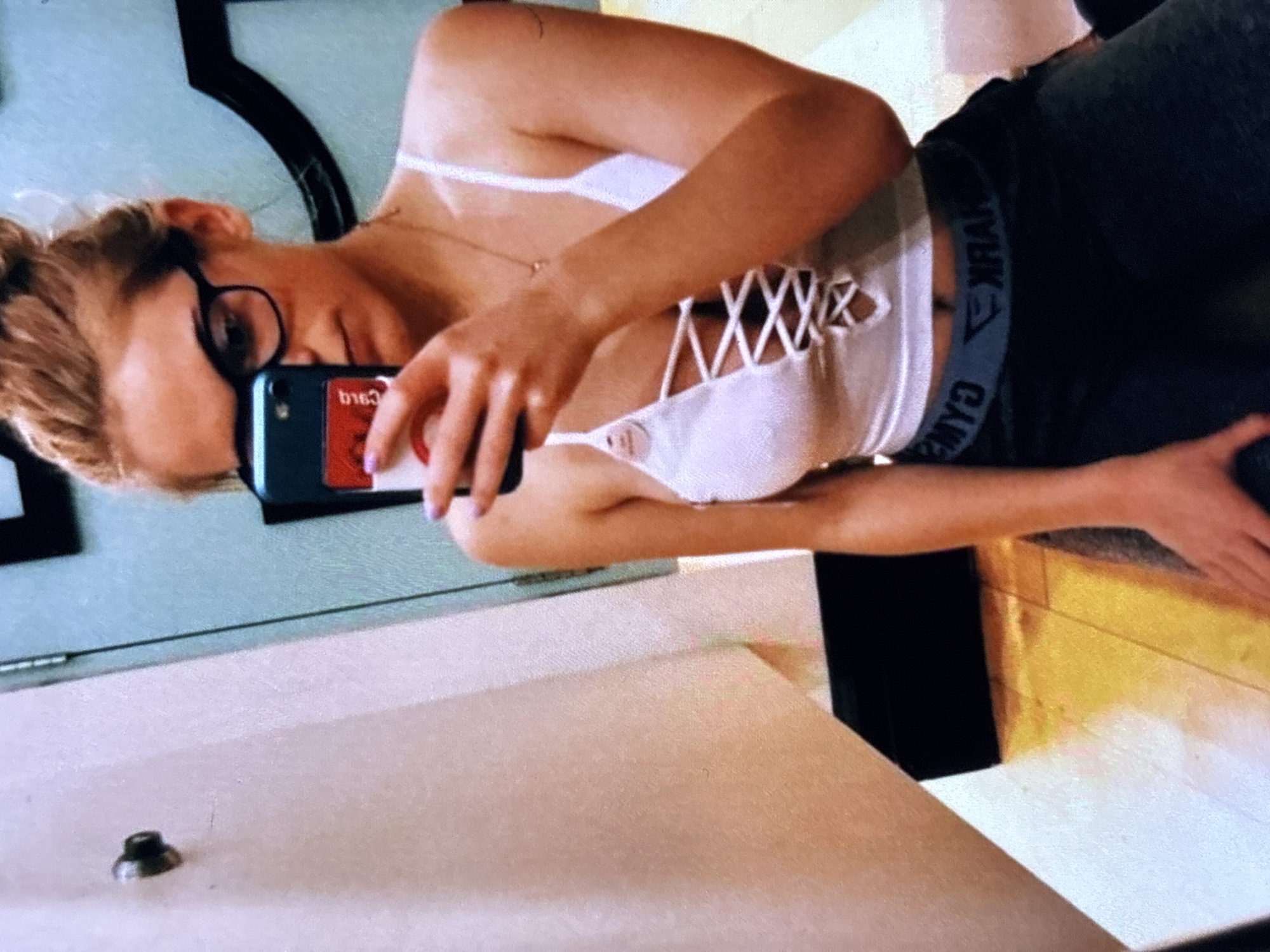 A young Caucasian woman is taking a mirror selfie in what appears to be a fitting room. She has blonde hair pulled up in a messy bun, is wearing large black-rimmed glasses, and has a white lace-up crop top on with gray athletic pants. Her left hand holds a smartphone with a red sticker on the back, and she is wearing a necklace. The background shows a light-colored wall and a door hinge, with a black bench or cabinet visible in the lower section of the image.