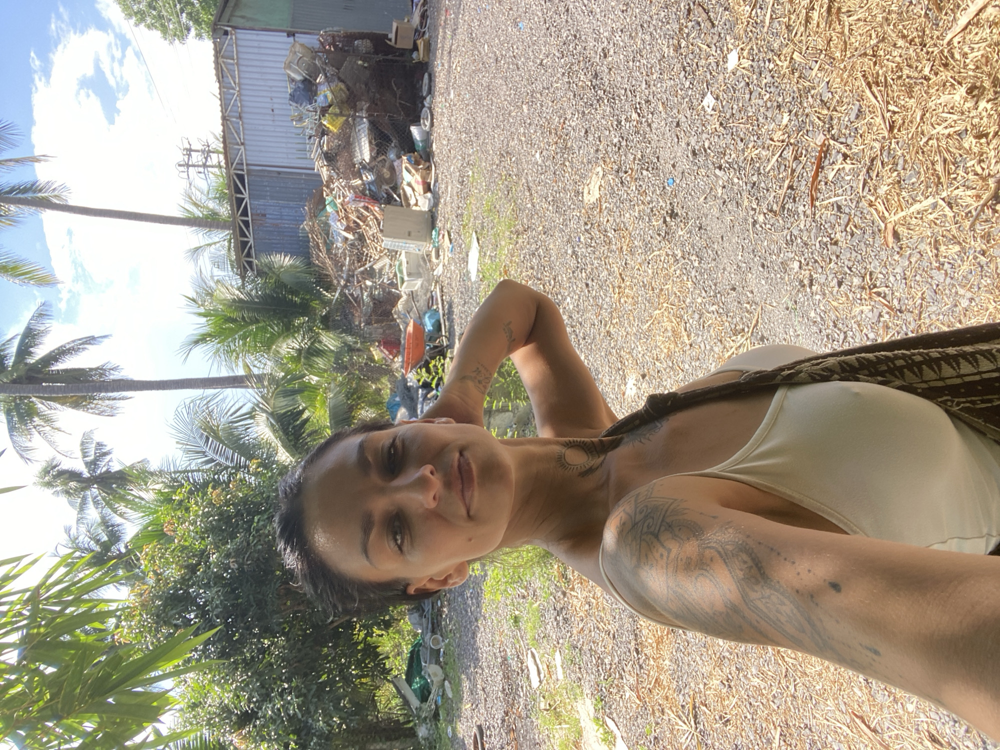 A woman stands in a sunny, outdoor area with a pile of discarded items in the background, located in Mũi Né, Vietnam. She is looking towards the camera, her head slightly tilted, with her right arm raised and hand behind her head. She has dark hair pulled back and tattoos on her left arm and neck. She is wearing a white tank top and a patterned bag is slung over her right shoulder. The ground is covered with gravel and dry vegetation. In the background, there is a corrugated metal building and a large accumulation of various trash and debris, including what appears to be furniture, plastic, and metal scraps. Palm trees and other greenery are visible around the scene.