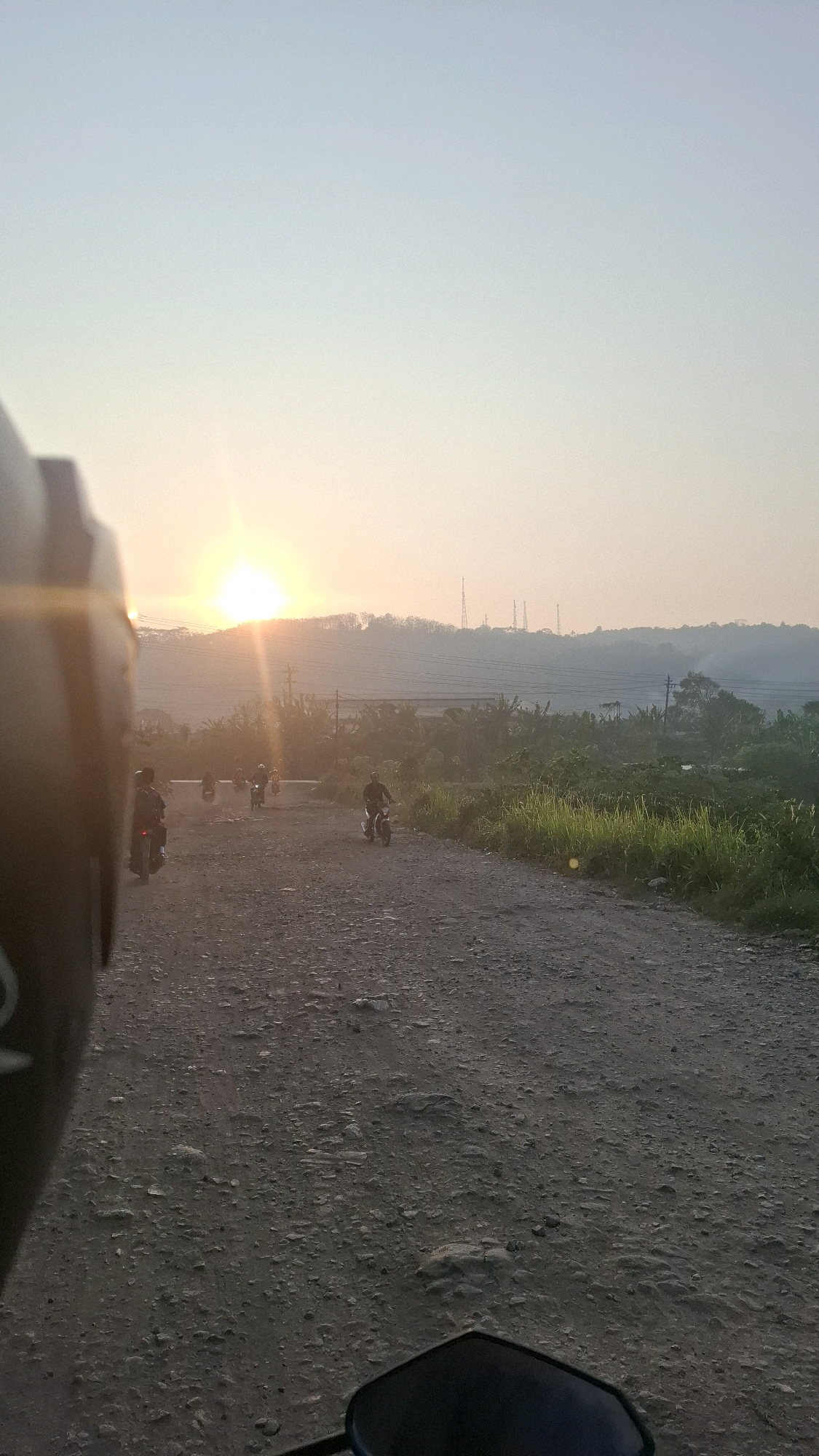 The image captures a scenic view of a sunset over a dirt road in Bandar Lampung, Indonesia. The sun, a brilliant orb, is low on the horizon, casting a warm glow across the landscape. The sky is a gradient of pale blue and soft orange, transitioning into a hazy yellow near the sun. Silhouetted against the sunset are rolling hills covered in vegetation, with several radio towers visible on the highest peak.
A dirt road, rough with stones and gravel, stretches into the distance, serving as the primary pathway in the scene. Several individuals on motorcycles are present on the road, some appearing to be traveling together. In the foreground, a helmet and a portion of a motorcycle are visible, suggesting the photo was taken from the perspective of someone riding a motorcycle. The vegetation along the sides of the road is lush and green, with tall grass and trees forming a natural border. The overall atmosphere is peaceful and serene, indicative of a rural setting within Bandar Lampung.