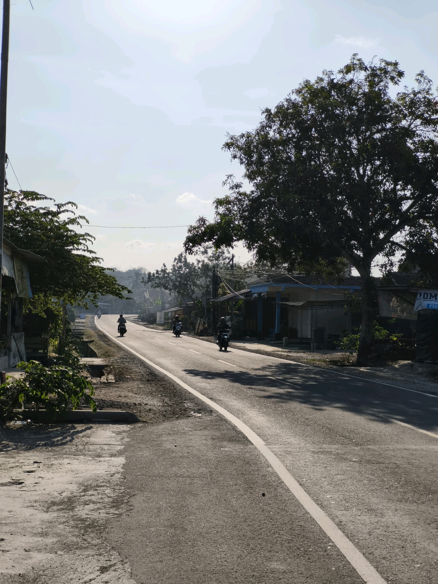 The image depicts a roadside scene in Cibuluh, Indonesia. The road, paved with asphalt, curves gently to the left, with a white line marking the center. On the right side of the road, there are several low-rise buildings with blue roofs, some appearing to be shops or homes. A large tree with dense foliage casts a shadow onto the road and the buildings. On the left side of the road, there is a sidewalk with vegetation and some dilapidated structures. Several motorcycles are visible on the road. One motorcycle with a rider is in the foreground on the right side, moving away from the viewer. Another motorcycle with a rider is in the middle ground on the left side, also moving away. Further in the distance, at least two more motorcycles are seen on the road. The sky is bright and partially cloudy, suggesting it is daytime. The overall impression is a typical rural or semi-urban street in Indonesia.