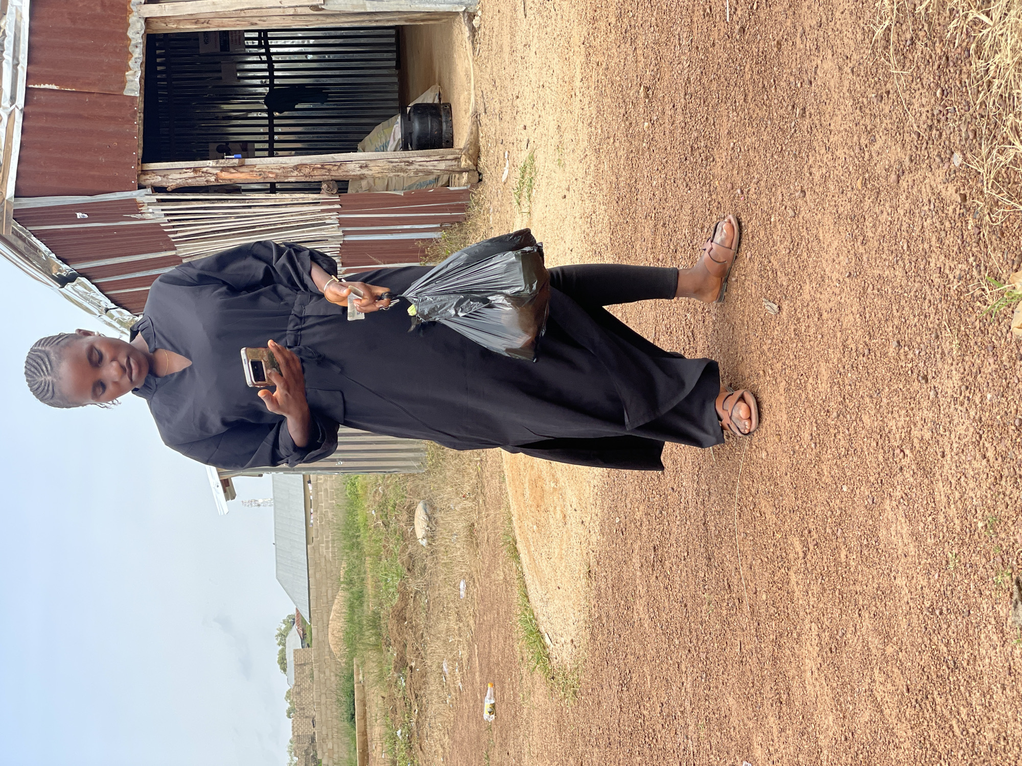 A young African woman stands outdoors, looking at her mobile phone. She is wearing a long black dress and black leggings, with brown sandals. In her right hand, she holds a black plastic bag containing items. The ground is covered in reddish-brown dirt and small stones. Behind her, there is a simple, rustic building with a corrugated metal roof and walls, appearing to be a small shop or dwelling. In the background, there are more buildings under construction and some sparse vegetation under an overcast sky. The scene is consistent with a rural or semi-urban environment in Durumi, Nigeria.