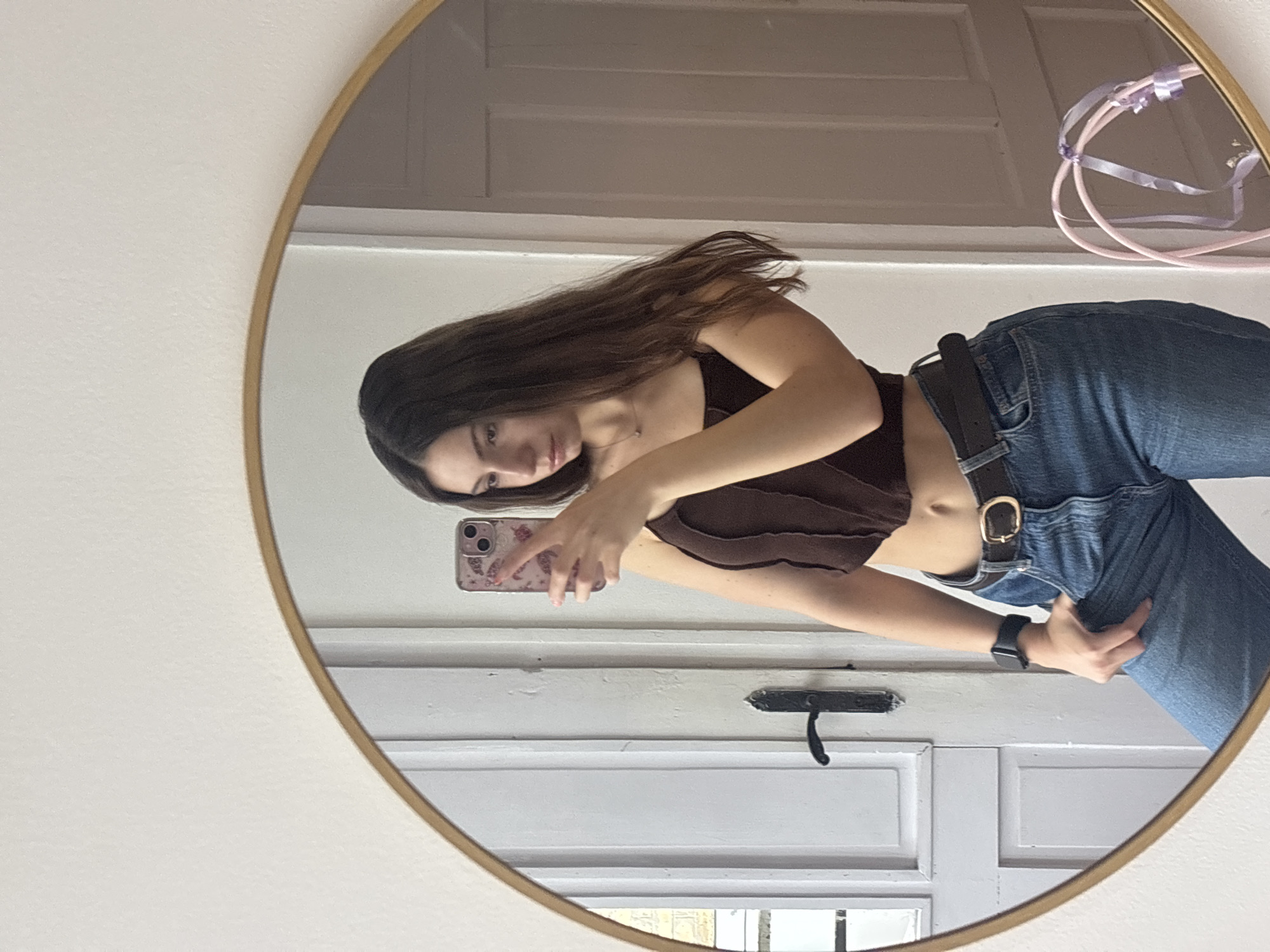 A young woman is taking a selfie in a round mirror. She is wearing a brown top and blue jeans with a black belt. She is holding a phone with a floral case. The background shows a white door with a black handle. The scene likely takes place indoors, possibly in a home. The location cannot be determined from the image.