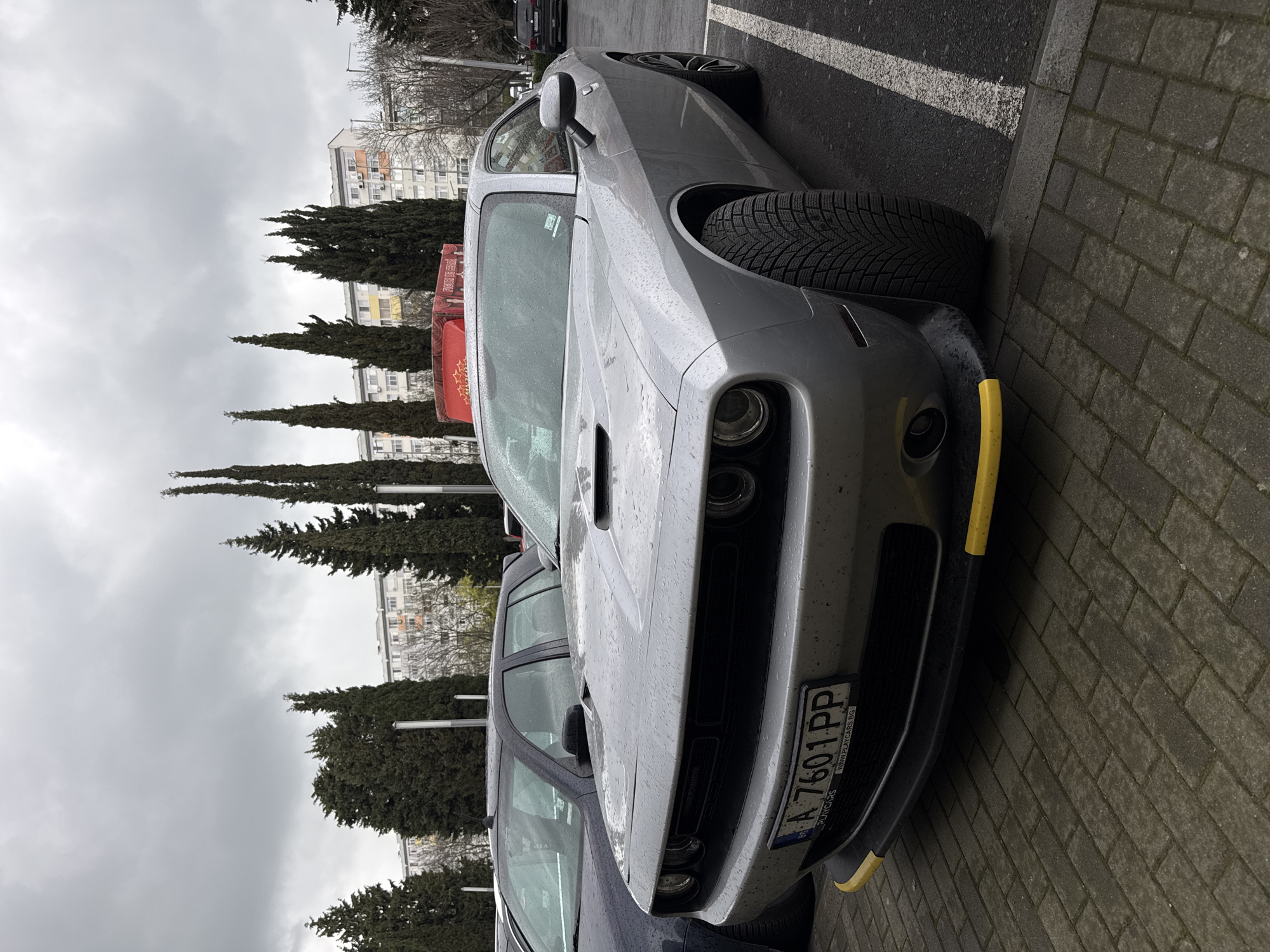 The image shows a silver Dodge Challenger parked on a brick sidewalk in Burgas, Bulgaria. The car is wet, suggesting recent rain. The license plate reads "A 7601 PP" and includes the website "WWW.PLAYCARS.BG". The front of the car has yellow splitter guards. The background includes evergreen trees, a multi-story building, and a gray, overcast sky. Another car is partially visible next to the Challenger. The scene appears to be an urban setting with typical European architecture in the background.