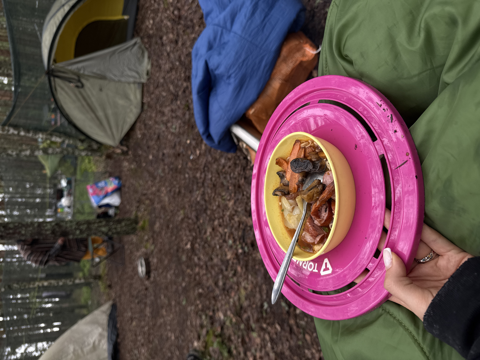 The image depicts a campsite scene, presumably in a wooded area near Luzhki, Russia. A person is holding a bright pink plate with a yellow bowl containing what appears to be a stew or soup made up of sausage, carrots, and mushrooms. A spoon is inserted into the bowl. The person is wearing a green jacket.
In the background, a partially open tent is visible, along with a blue blanket and a brown bag. Further in the background, camping gear and a chair are visible. The ground is covered with fallen leaves and dirt, and trees are scattered throughout.