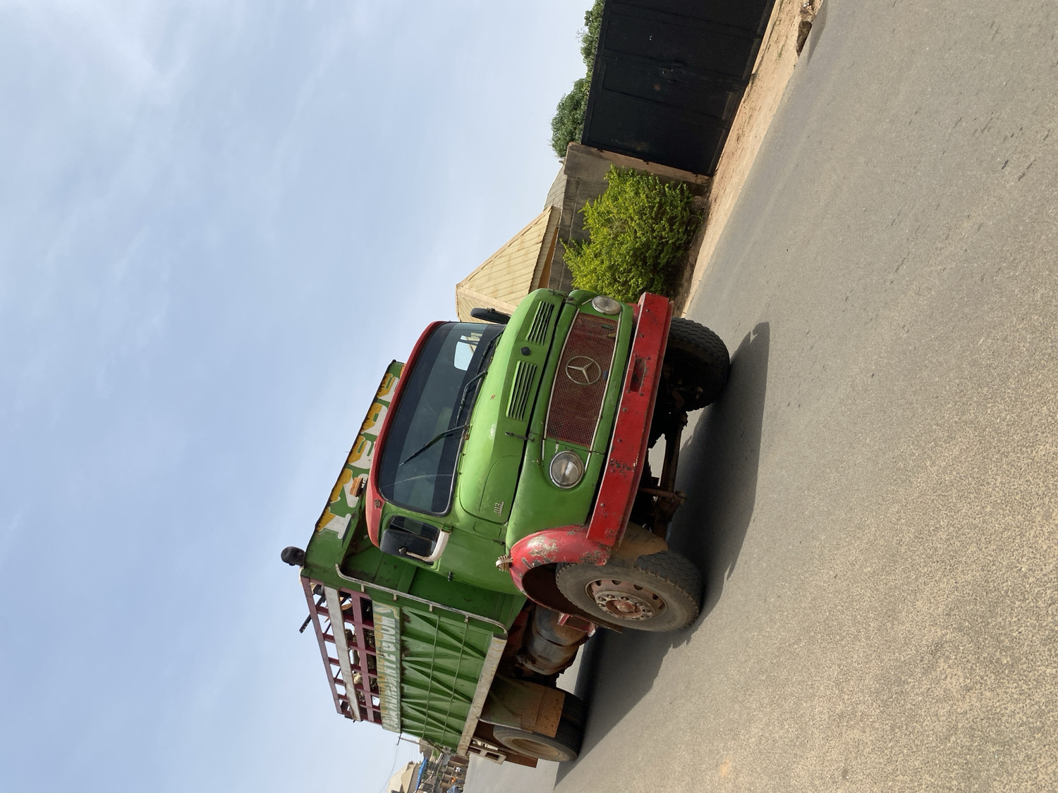 A robust, older model Mercedes-Benz truck, likely a cargo or dump truck, is the main subject of the image, captured from a slightly elevated, side-front perspective. The truck features a faded green cab and cargo bed, contrasted with a bright red front bumper and red trim along the roofline. The grille, showing signs of wear and rust, prominently displays the iconic Mercedes-Benz star logo. The vehicle's overall appearance suggests significant use and durability.
The truck is situated on a paved road, which extends across the foreground and to the right side of the frame. In the background, to the right, a dark, possibly black, metal gate is visible behind a low wall, accompanied by green bushes or foliage. The sky is clear and light blue, indicating bright daylight conditions, with the strong sunlight casting distinct shadows from the truck onto the road.
Visible text on the side of the truck's green cargo bed includes "WOOD STORE" in white lettering on a yellow band. Below this, another line of text is partially legible, appearing to contain "BARAKIN AHAMATE GOM" or similar wording, suggesting a local business or origin, consistent with the Barakin Akawo, Nigeria context. The scene portrays a functional vehicle in a daily street setting under clear weather conditions.