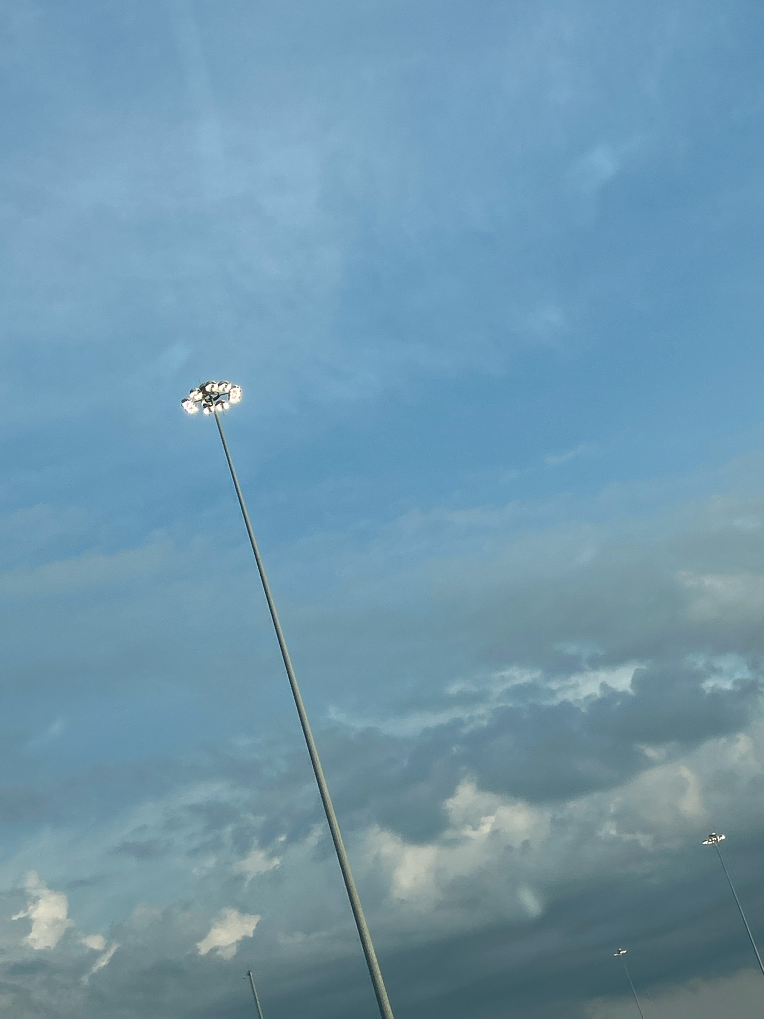 A tall, thin pole topped with a cluster of bright lights extends upwards into a cloudy blue sky. The pole is positioned off-center to the left of the frame, reaching diagonally upwards and to the right. The lights at the top are illuminated, creating a halo effect against the sky. In the distance, two smaller poles with similar lights are visible, also against the cloudy backdrop. The sky is filled with a mix of light and dark clouds, suggesting it might be overcast or late in the day. The overall scene captures an outdoor environment, possibly a sports field or stadium, in Robinson Township, USA.