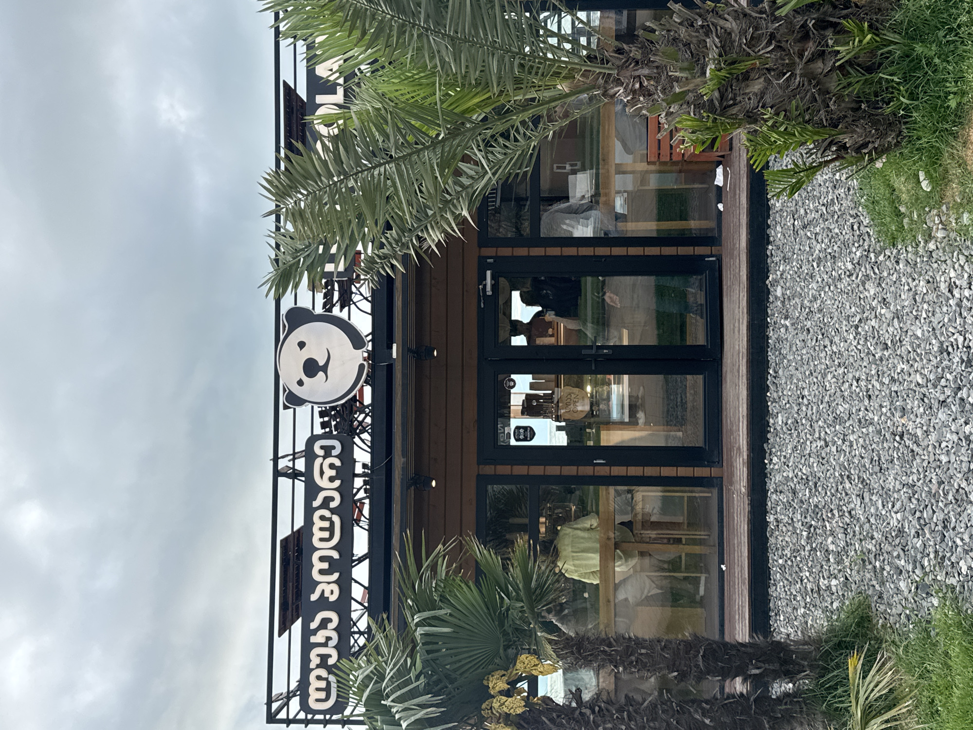 The image shows the front of a building with a sign featuring a panda bear logo and Georgian text. The building has a brown wooden facade with a black-framed glass entrance, through which one can see a person inside. Palm trees flank the entrance on either side. A gravel ground covers the area around the building, with grass growing along the edge. The sky is overcast. This is in Batumi, Georgia.