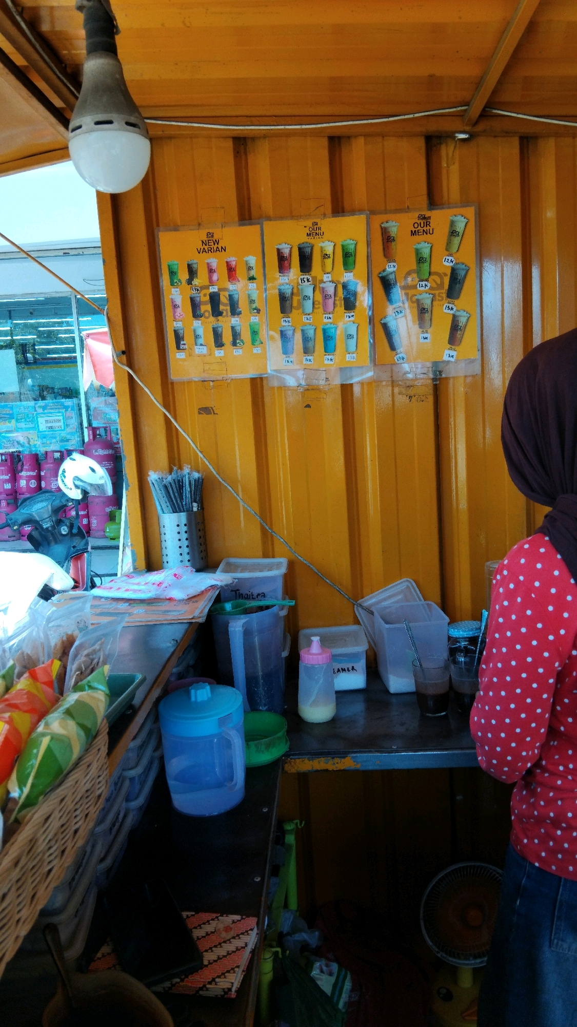 The image depicts a street food stall in Watesalit, Indonesia, with a focus on drinks. The stall is constructed from corrugated yellow metal. A hanging LED bulb provides illumination. Two framed menus displaying various drinks are mounted on the stall's wall, showcasing colorful beverages in plastic cups with prices such as "15K" and "12K." Below the menus, a collection of plastic cups, straws, and various containers are visible on a dark countertop. These include a large blue pitcher, a green bowl, and smaller containers labeled "Thaitra" and "LAMER." A basket filled with snacks, possibly wrapped in colorful plastic, sits on the left side of the counter. In the background, to the left, a motorcycle with a white helmet is parked, and pink gas cylinders are stacked. On the right side, a person wearing a red and white polka-dotted shirt and a dark headscarf is partially visible, facing away from the camera and towards the stall. A small oscillating fan is situated on the lower right portion of the image. The overall impression is that of an active, outdoor beverage vendor.