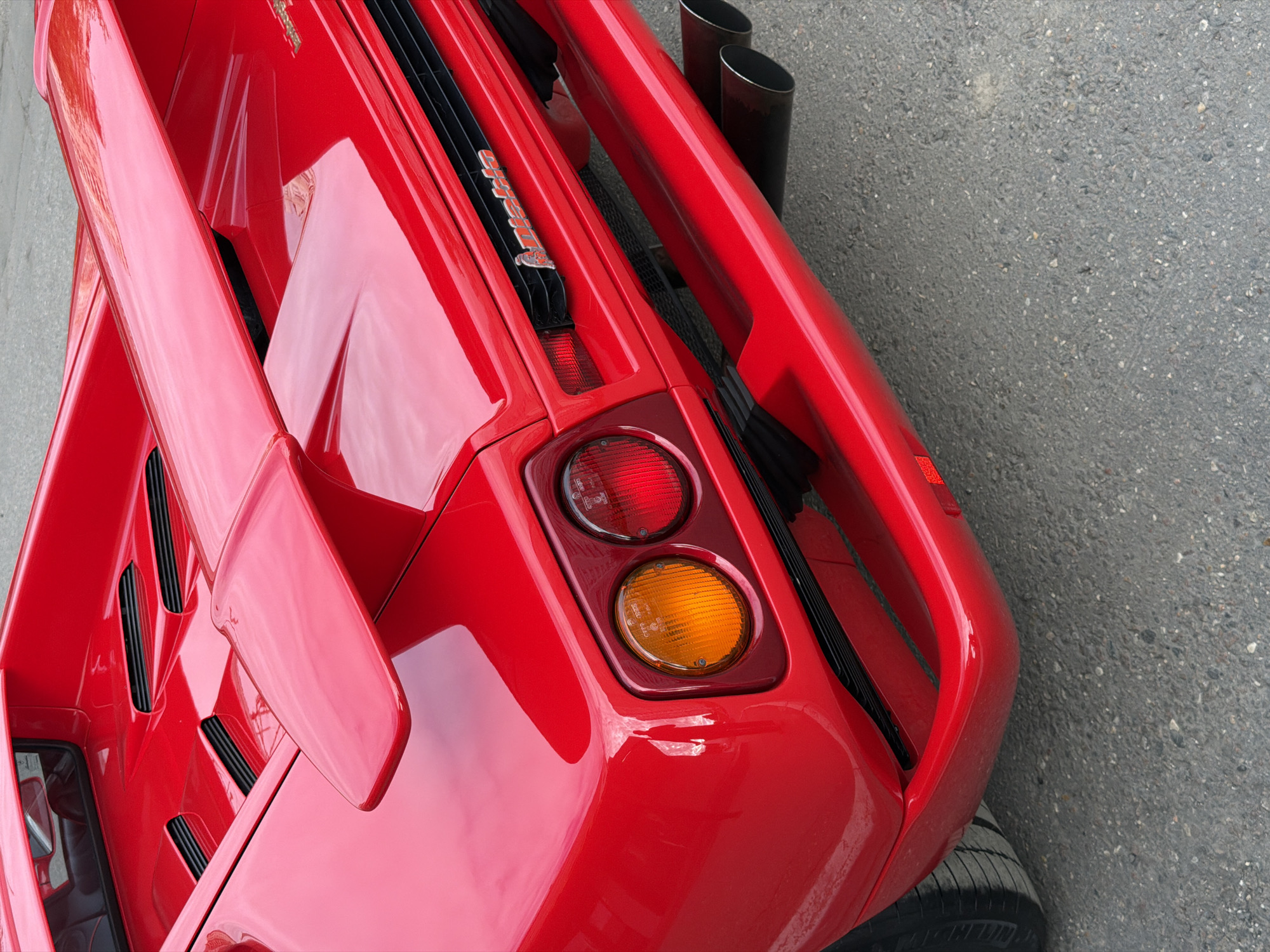 The image presents a high-angle, close-up view of the rear of a bright red Lamborghini Countach, parked outdoors on an asphalt surface. The car's distinct angular design and highly reflective red paintwork are prominent.
Visible on the upper right side of the car's rear are two large, metallic exhaust pipes. Below these, a black slatted engine cover spans the width of the vehicle, with the name "Countach" clearly inscribed in silver lettering on a black strip. The rear lights, located on the lower left, feature two circular lenses: a red brake light on top and an amber turn signal below.
A significant red rear spoiler with a sharp, aerodynamic shape extends from the car's body. In the lower right corner, a segment of a black tire is visible, contrasting with the grey, textured asphalt. The scene is well-lit by natural light, suggesting it is daytime with clear weather conditions. The car is stationary, and no individuals or specific activities are depicted. The provided context indicates the location is Moscow, Russia.