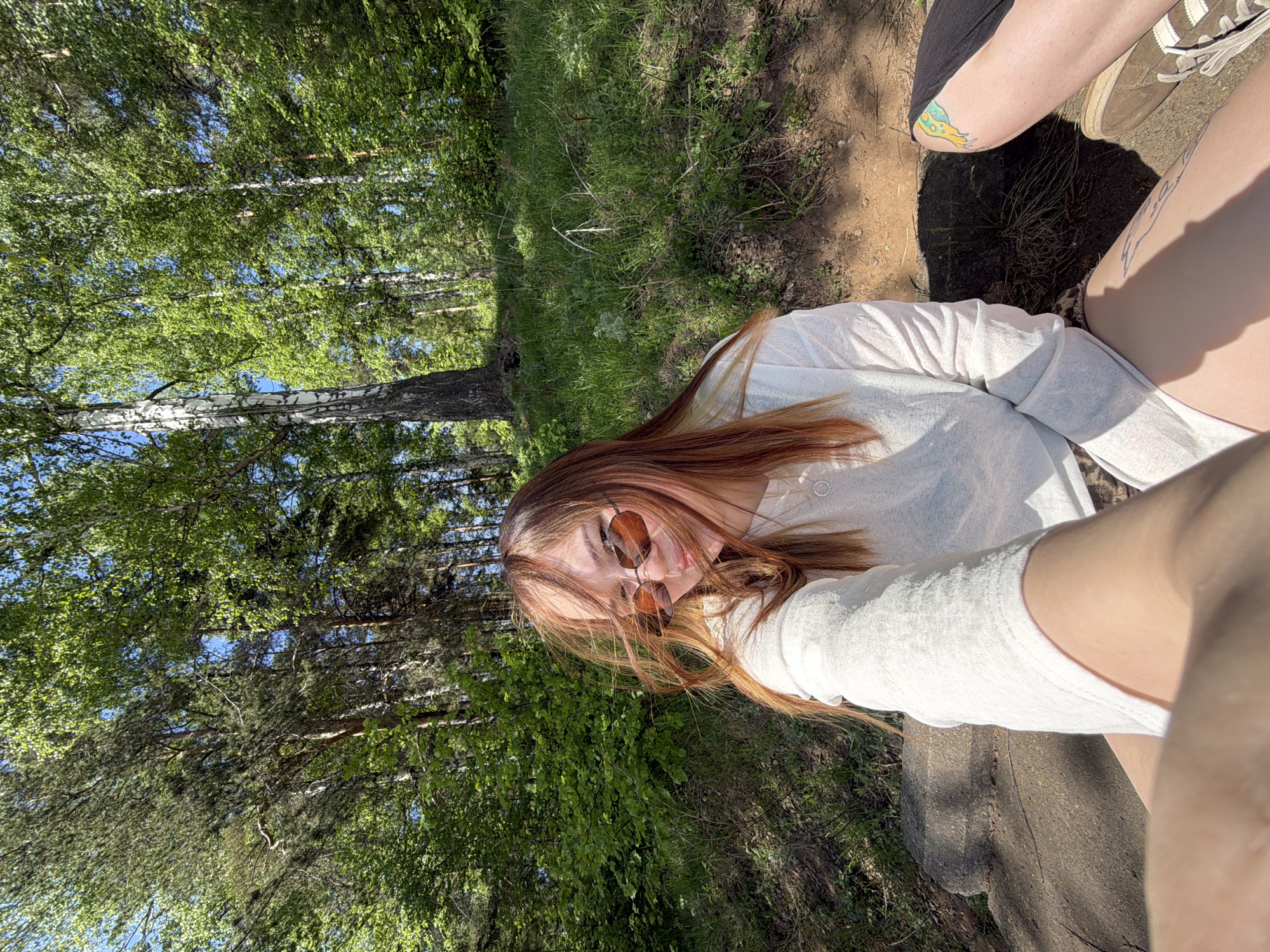 A young woman with light brown hair and wearing sunglasses is sitting in a grassy, wooded area, possibly in a park. She is wearing a light-colored top. Two pairs of legs are visible, suggesting there is at least one other person present. One pair of legs has a tattoo and wears tennis shoes. The forest is in the background. The location may be in the city of Chaikovsky, Russia, as requested.