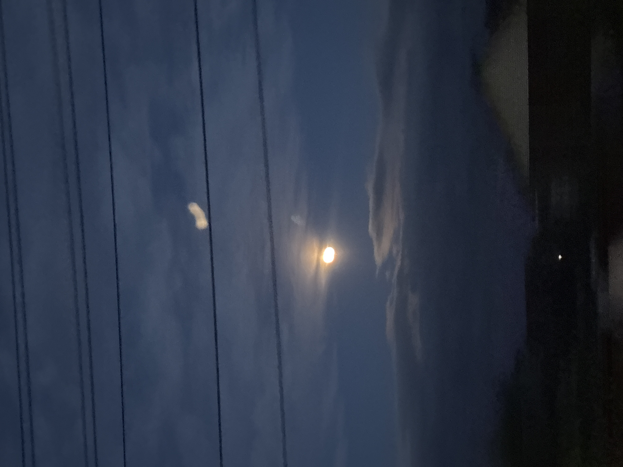 The image shows a night scene in what appears to be an urban or suburban environment, likely in Chaikovsky, Russia, due to the prompt's mention. The sky is a gradient of dark blue and grey, with visible clouds and the moon shining prominently. Power lines run vertically across the left side of the frame. A dark, indistinct structure or building is partially visible on the right side of the image. The scene is generally dimly lit, with the moon being the primary light source. No people are directly visible in the image.
