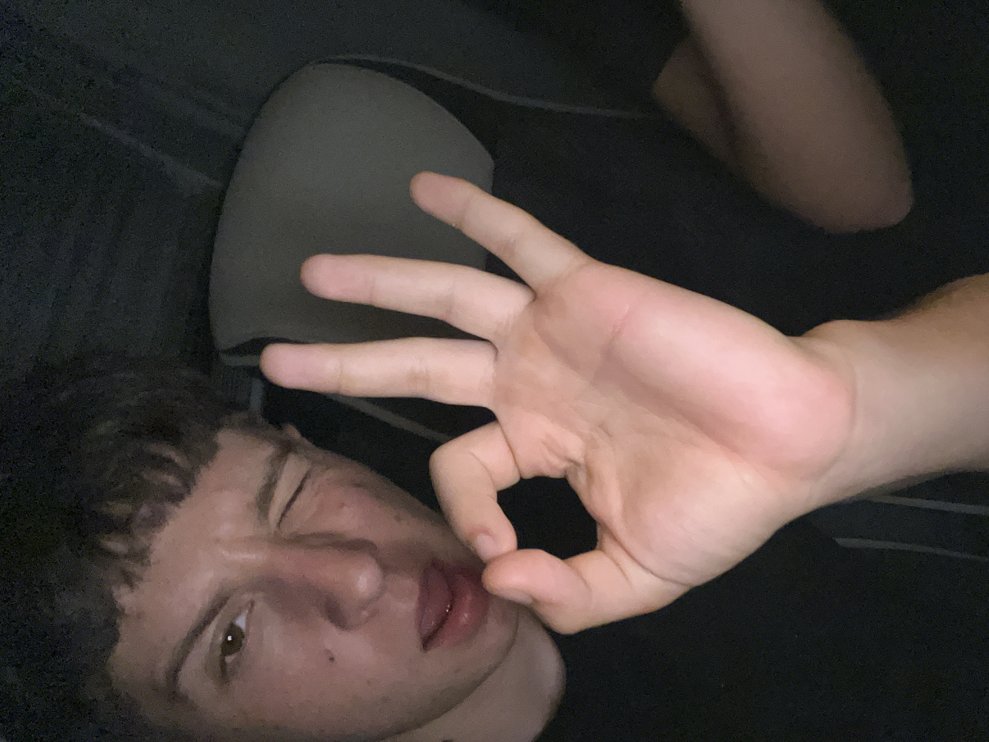 A young person is in a car, possibly at night. The person is making a circle shape with their thumb and forefinger, winking at the camera. The interior of the car is visible, including the seat headrest. The location is not Vinitsa, Bulgaria.