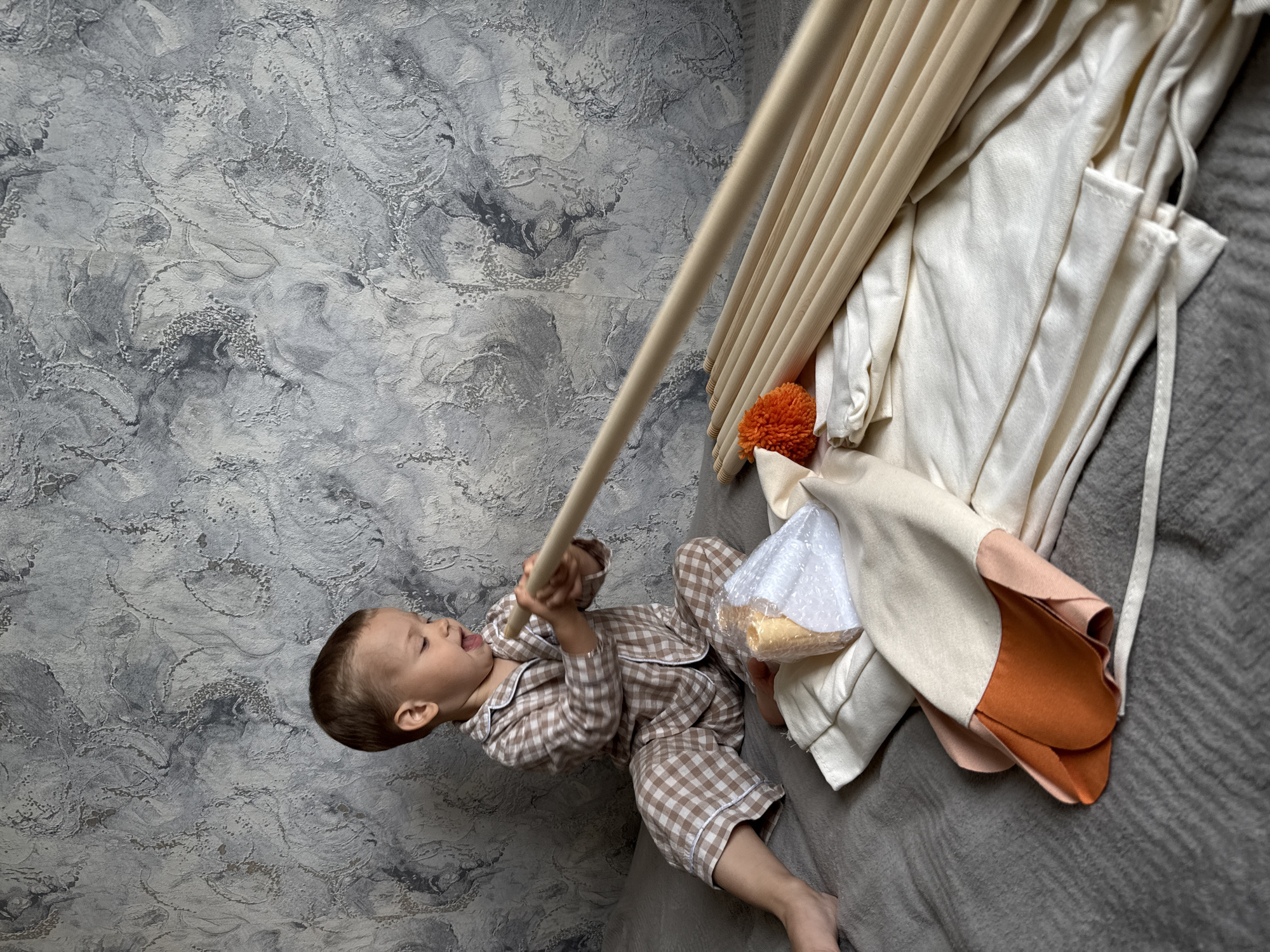 A young child in brown and white checkered pajamas is lying on a gray surface next to a partially assembled play tent. The child is holding one of the tent poles and appears to be either speaking or sticking out their tongue. Beside the child are the tent's fabric components, including white and tan sections, along with an orange pom-pom. The background is a gray, textured wall. The location is Rusanovo, Russia.