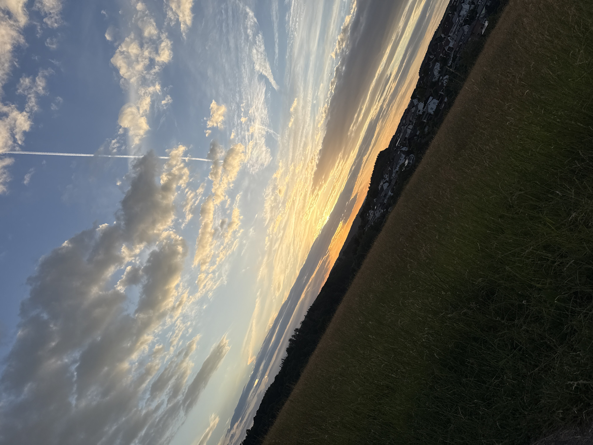 The image depicts a scenic view of the town of Oberquembach, Germany, during sunset. The foreground is dominated by a field of tall, dry grass that slopes downwards from left to right. In the midground, a dense line of trees forms the boundary between the field and a residential area. The houses in Oberquembach are clustered on the hillside, with their rooftops visible against the darkening landscape. The sky above is a canvas of dramatic colors, with bands of orange, yellow, and blue blending together as the sun sets. Wispy clouds are scattered across the sky, some illuminated by the fading sunlight, while others are darker and more ominous. A distinct contrail from an airplane stretches vertically across the upper portion of the sky, adding a modern element to the otherwise natural scene. The overall impression is one of a serene and picturesque rural evening.